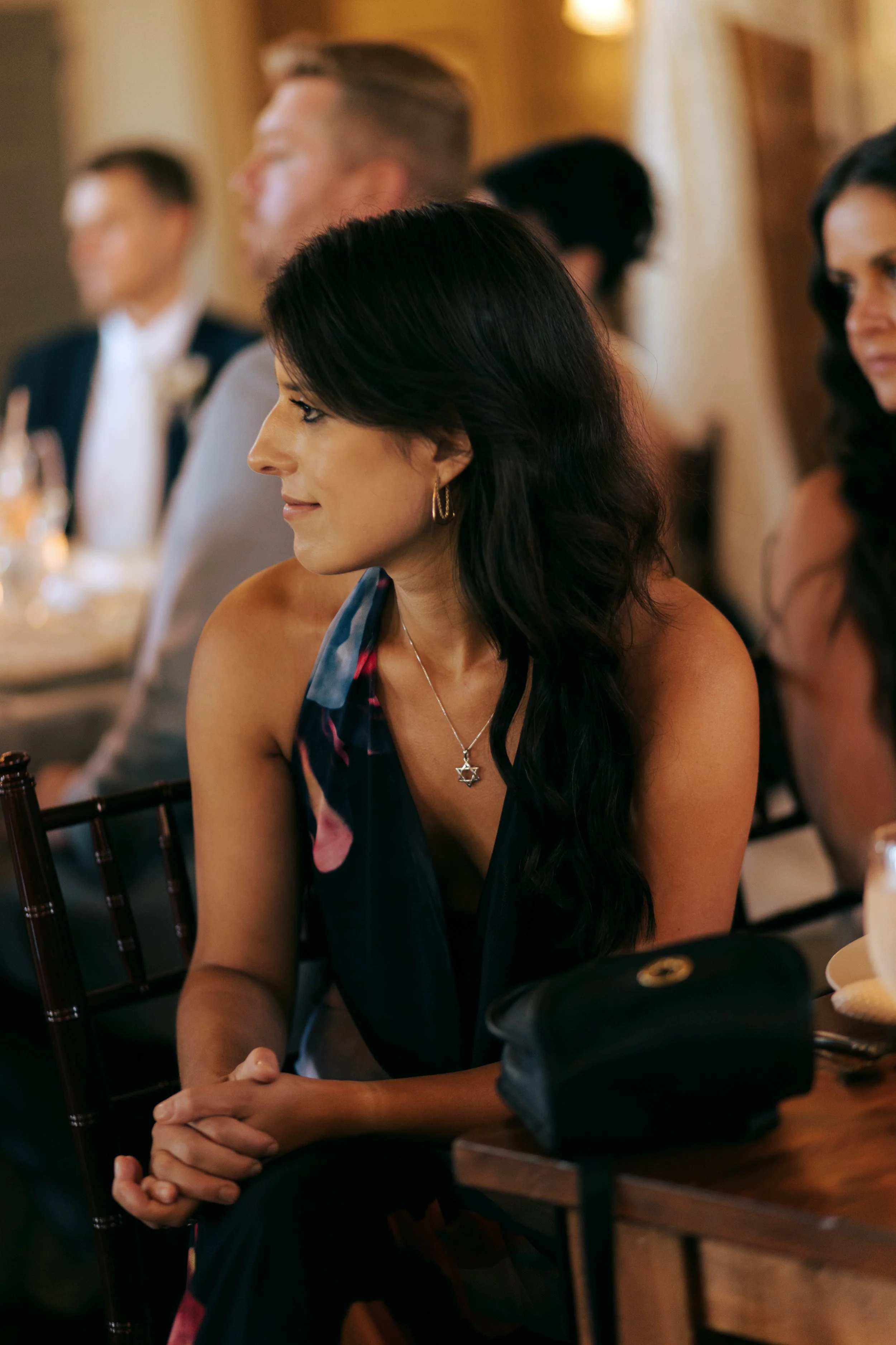 A woman with long dark hair, wearing a sleeveless floral dress, sitting at a formal dinner, wearing a Star of David necklace.