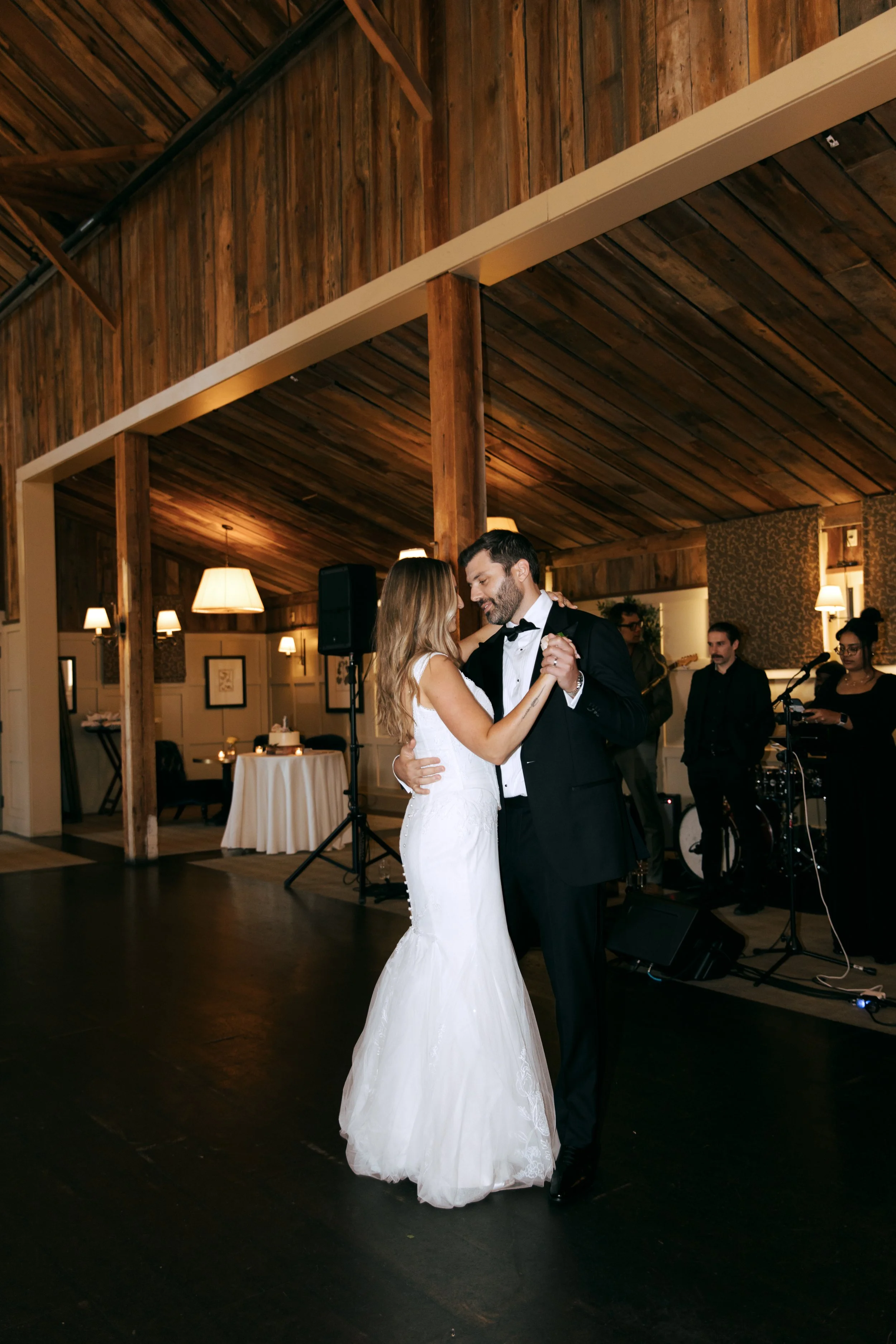 A bride and groom dancing in a rustic wooden wedding reception hall with a live band playing in the background.