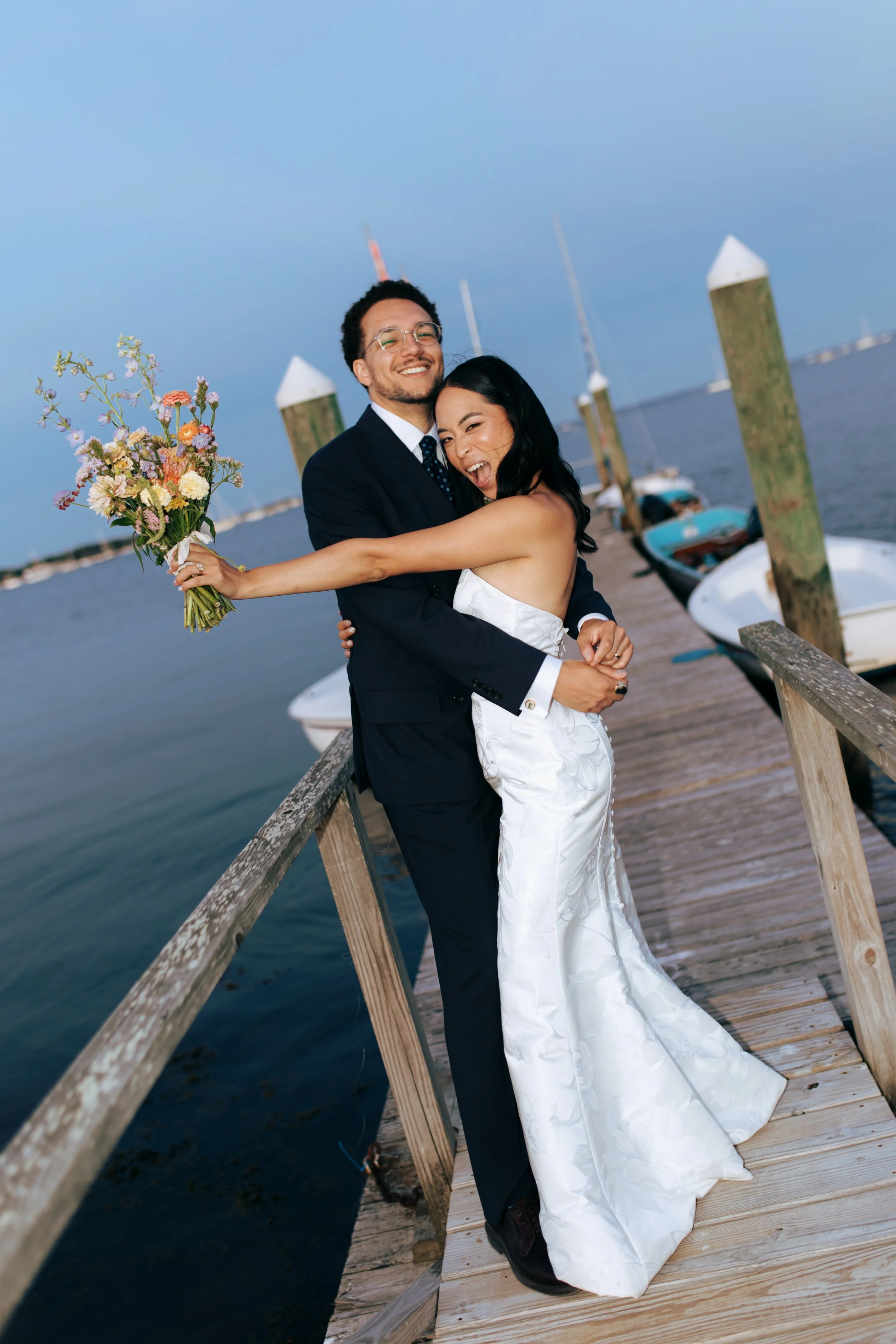 A happy couple in wedding attire embracing on a dock near the water, with boats and poles in the background.
