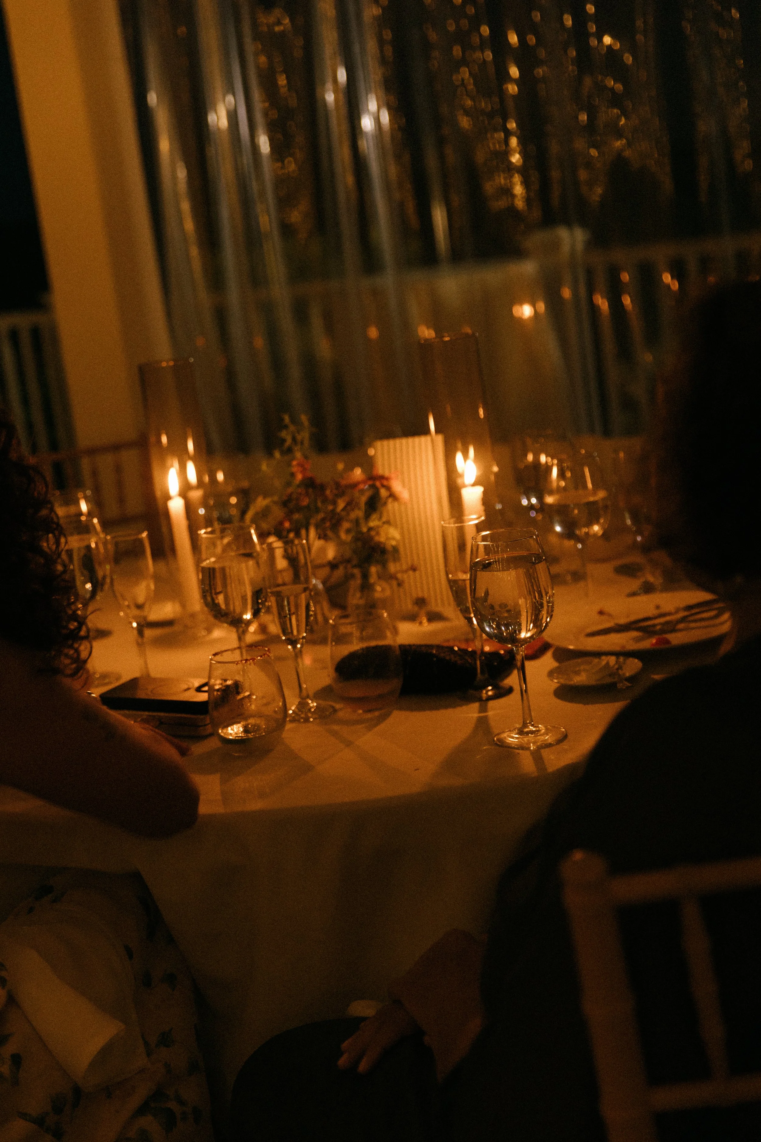 A dimly lit dinner table set for an event with multiple glasses of water, candles, and floral decorations, viewed in a warm, intimate ambiance.