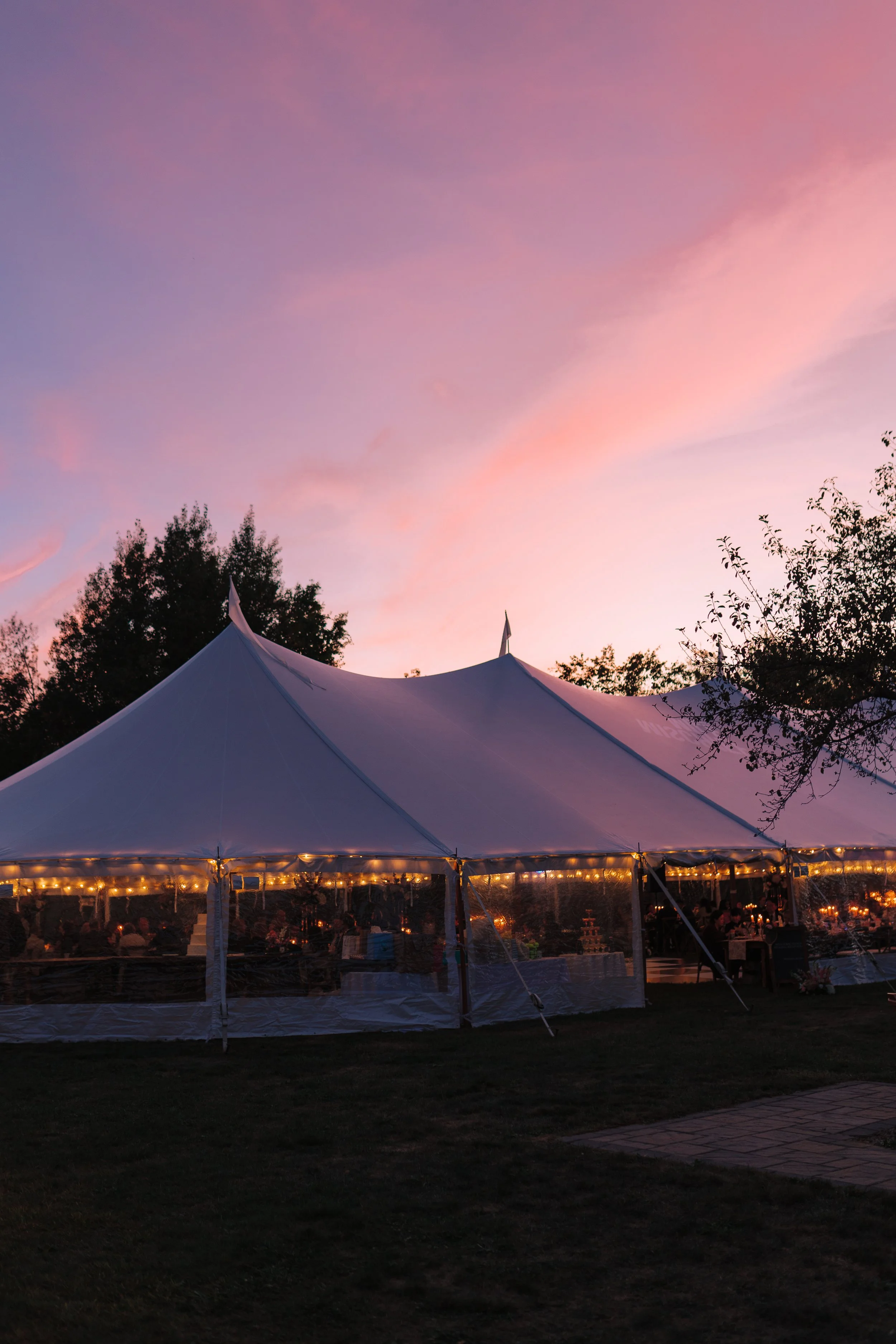 Outdoor event tent with string lights under a pink and purple sunset sky, surrounded by trees.