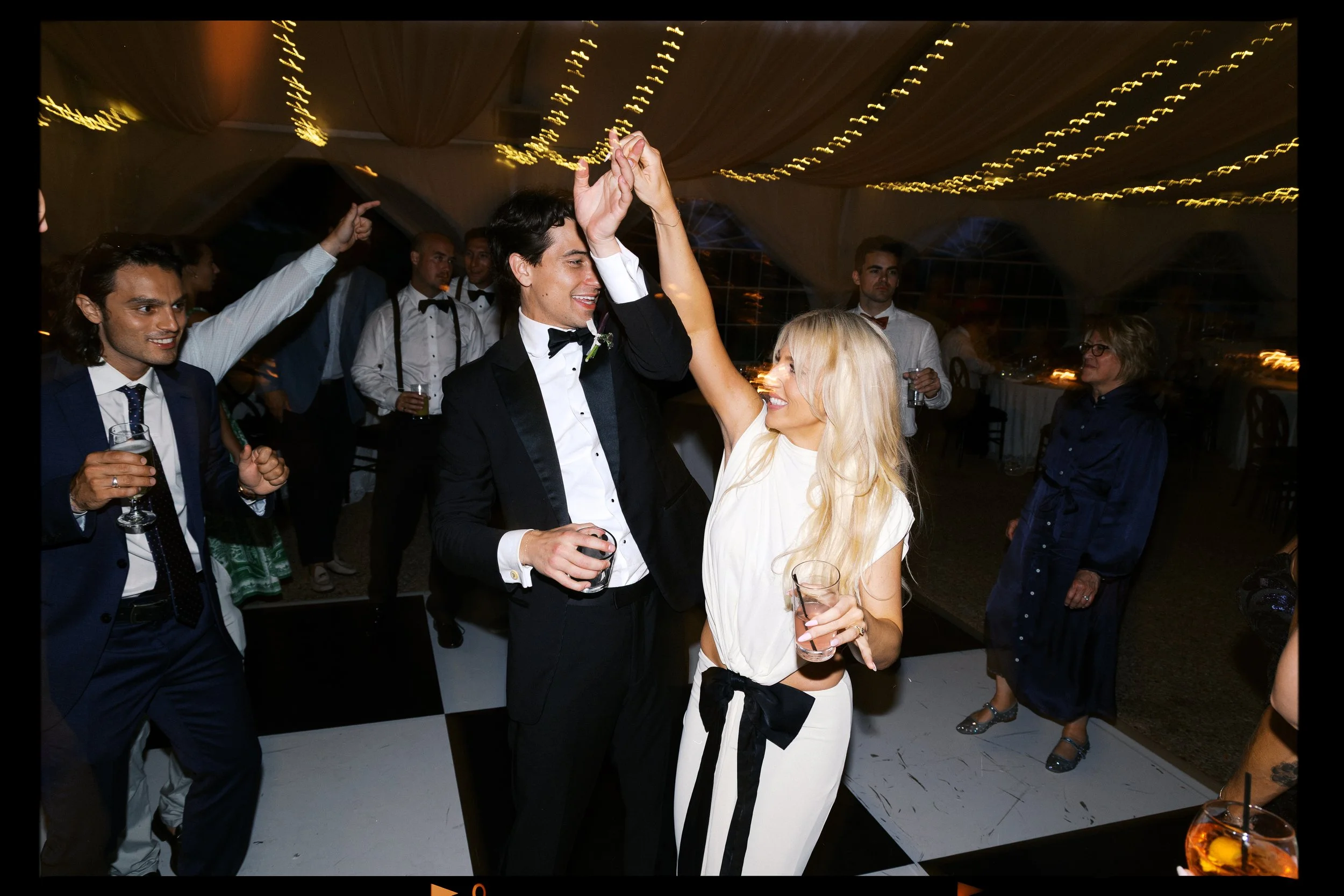 People dancing and celebrating at a wedding reception, with string lights overhead and a checkered dance floor.