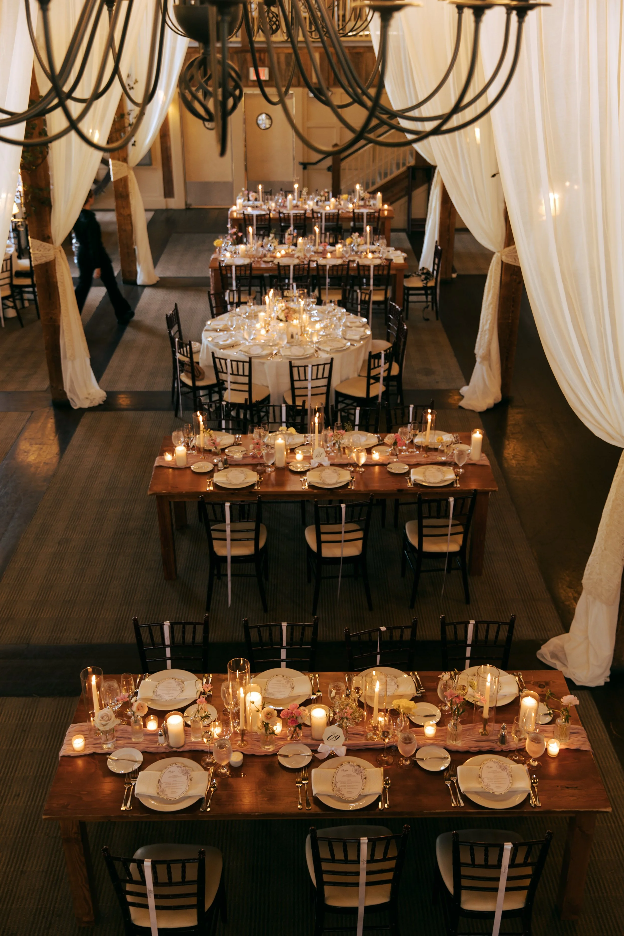 A top-down view of an elegant wedding or event reception with decorated tables, candles, flowers, and drapery in a large hall.