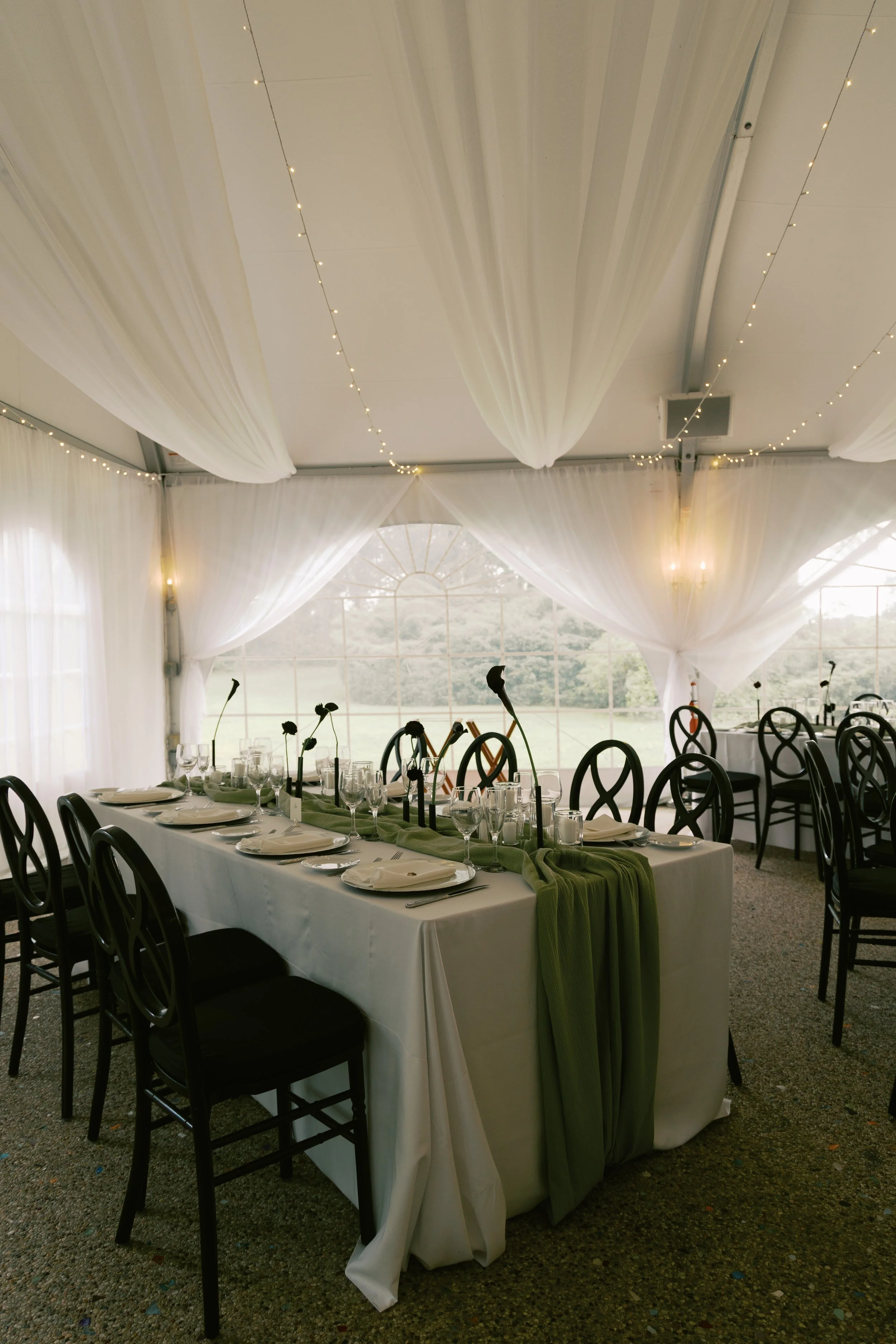 Elegant banquet table set for a special event under a white tent with draped fabric and string lights, decorated with black chairs, glassware, plates, and a green table runner with floral centerpieces.