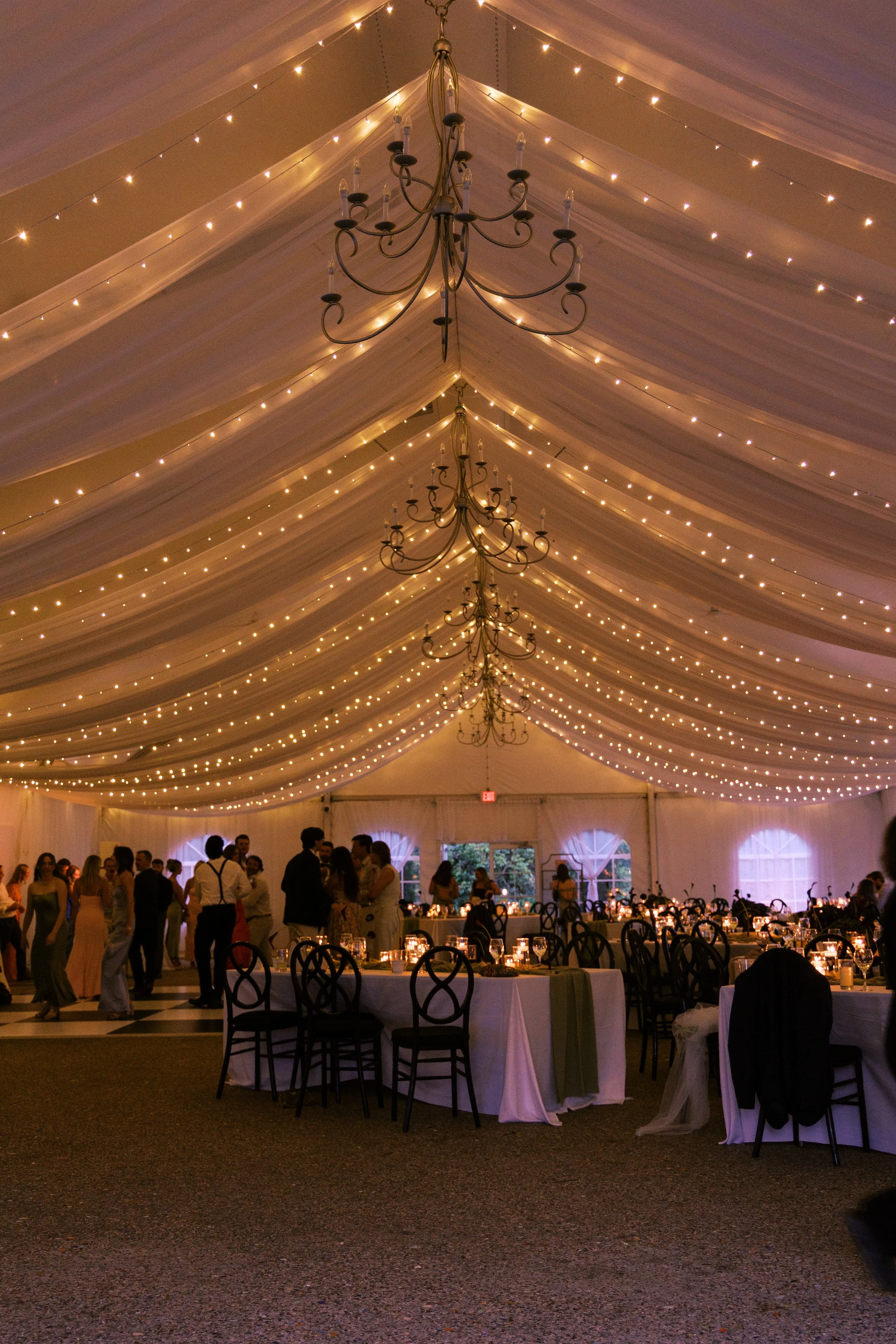 An elegant event space decorated with white drapery and string lights on the ceiling, with black chandeliers hanging down, and people gathered and dancing underneath.