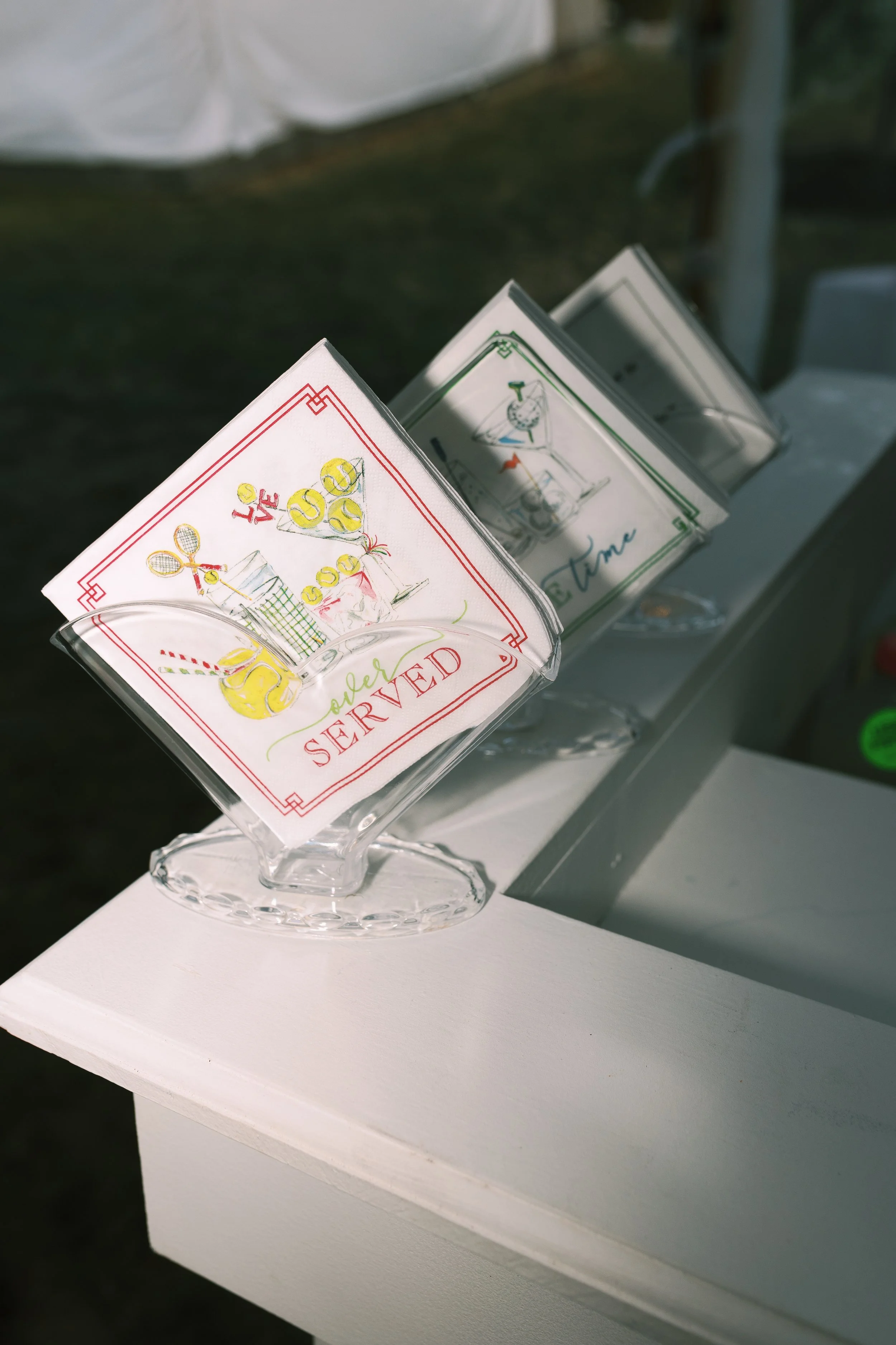 Napkins with colorful illustrations and text, placed on a glass napkin holder on a white table.