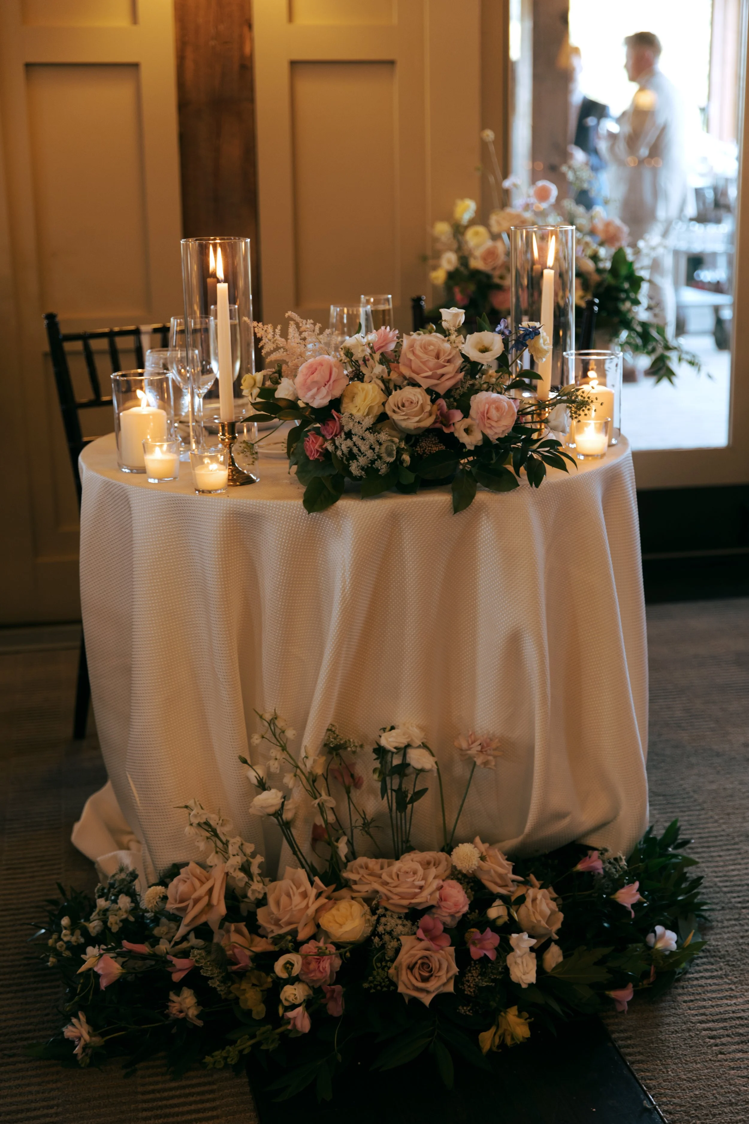 A round table with white tablecloth decorated with pink and white roses, lit candles, and glass vases, set for a wedding reception or special event, with a floral backdrop and a person in the background through a glass door.