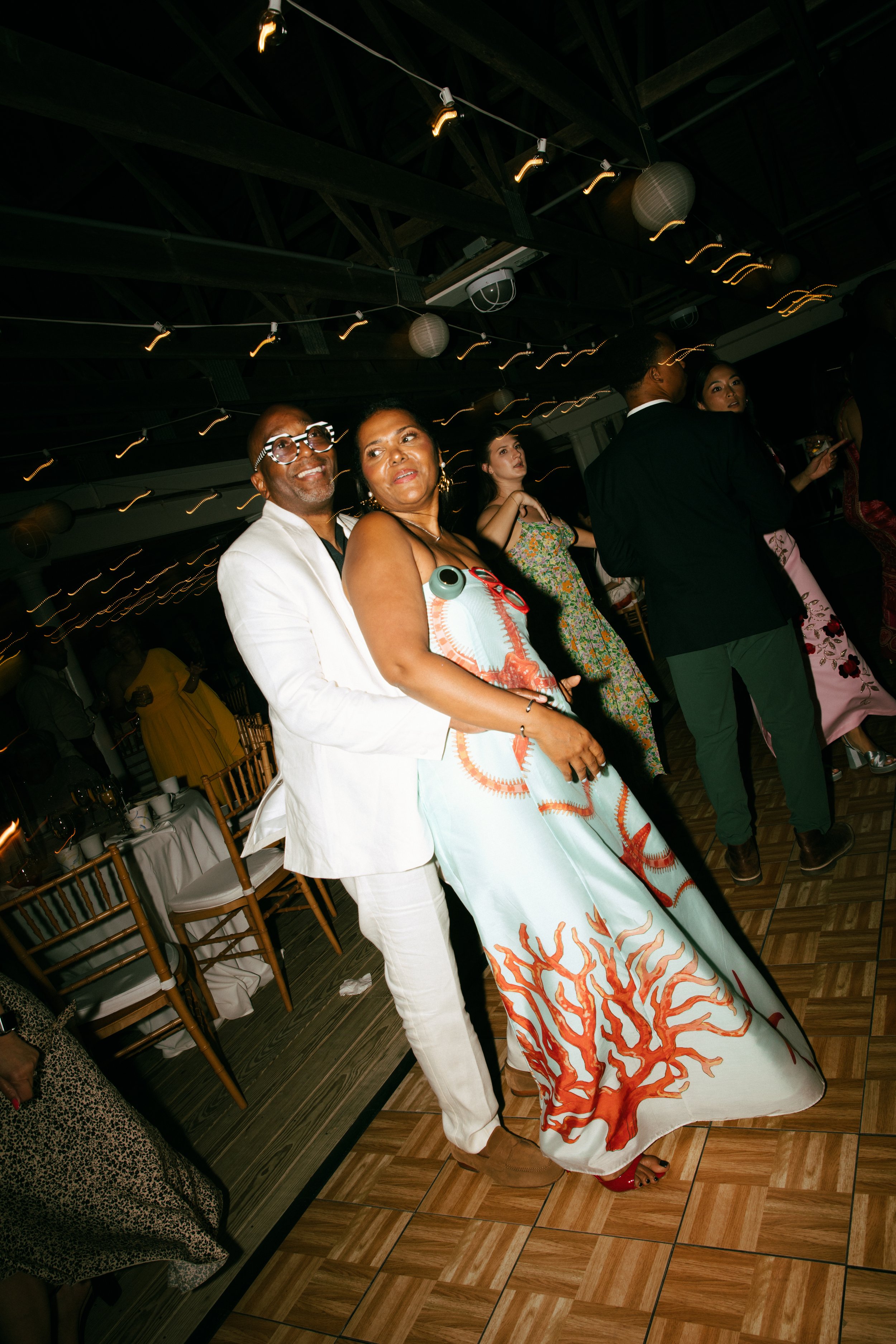 People dancing at a party, two individuals in the foreground smiling and posing for the camera; the man is in a white suit with glasses, and the woman is in a colorful dress with a coral reef design.