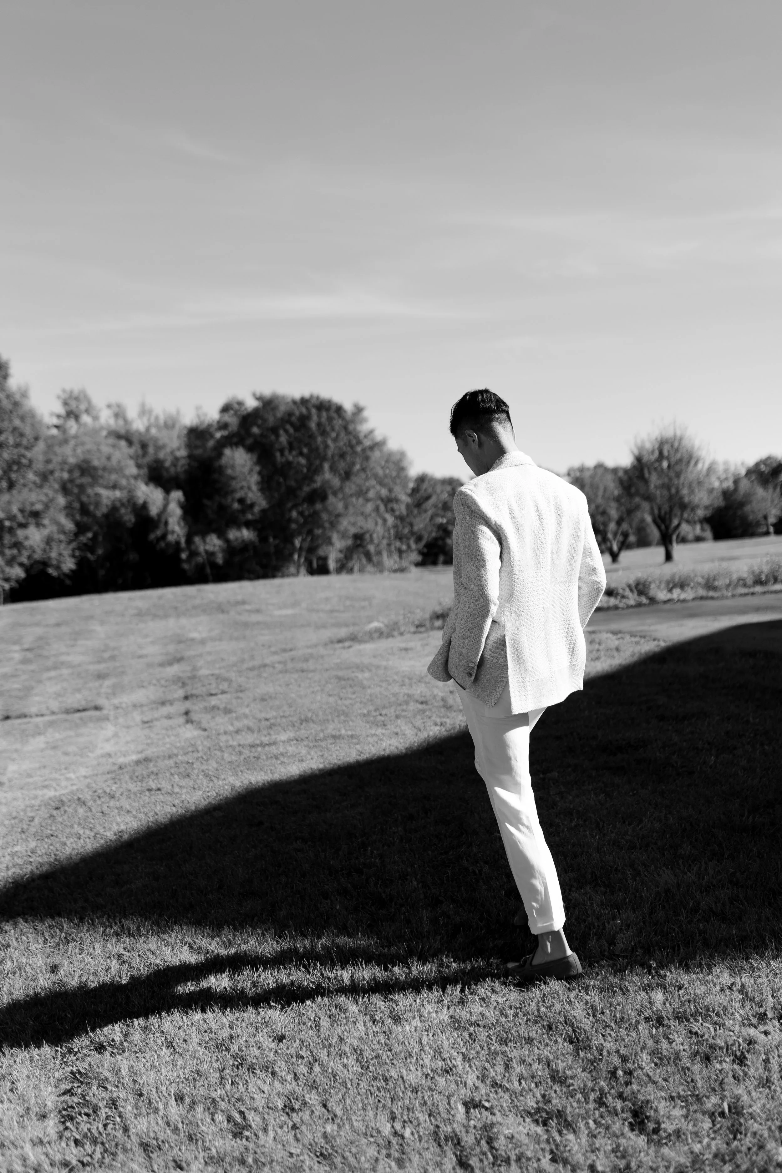 A man in formal attire walking on a grassy field with trees in the background, cast in black-and-white.