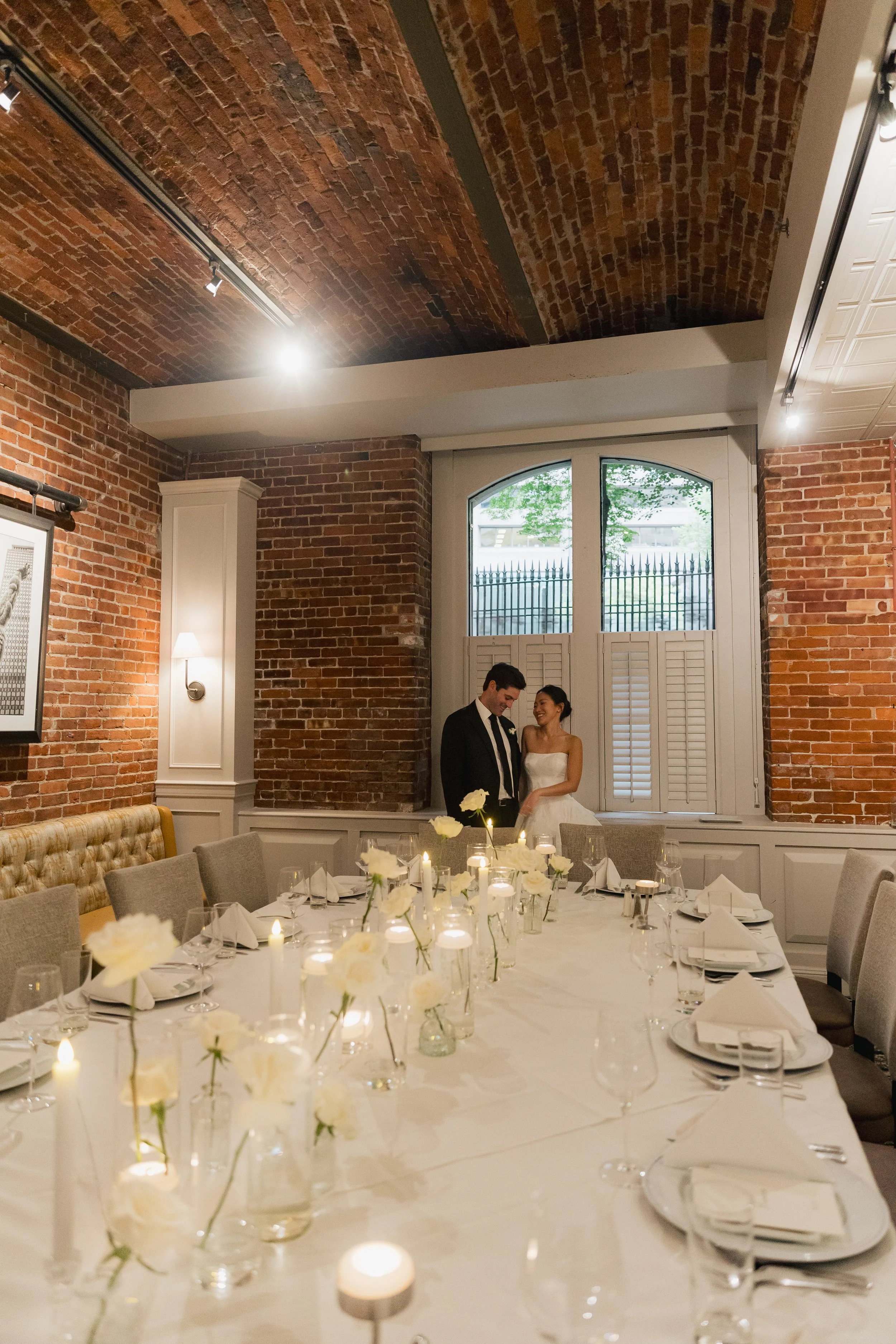A bride and groom standing close and smiling inside a decorated dining room with a long table set for a wedding celebration, brick walls, and a large window with shutters.