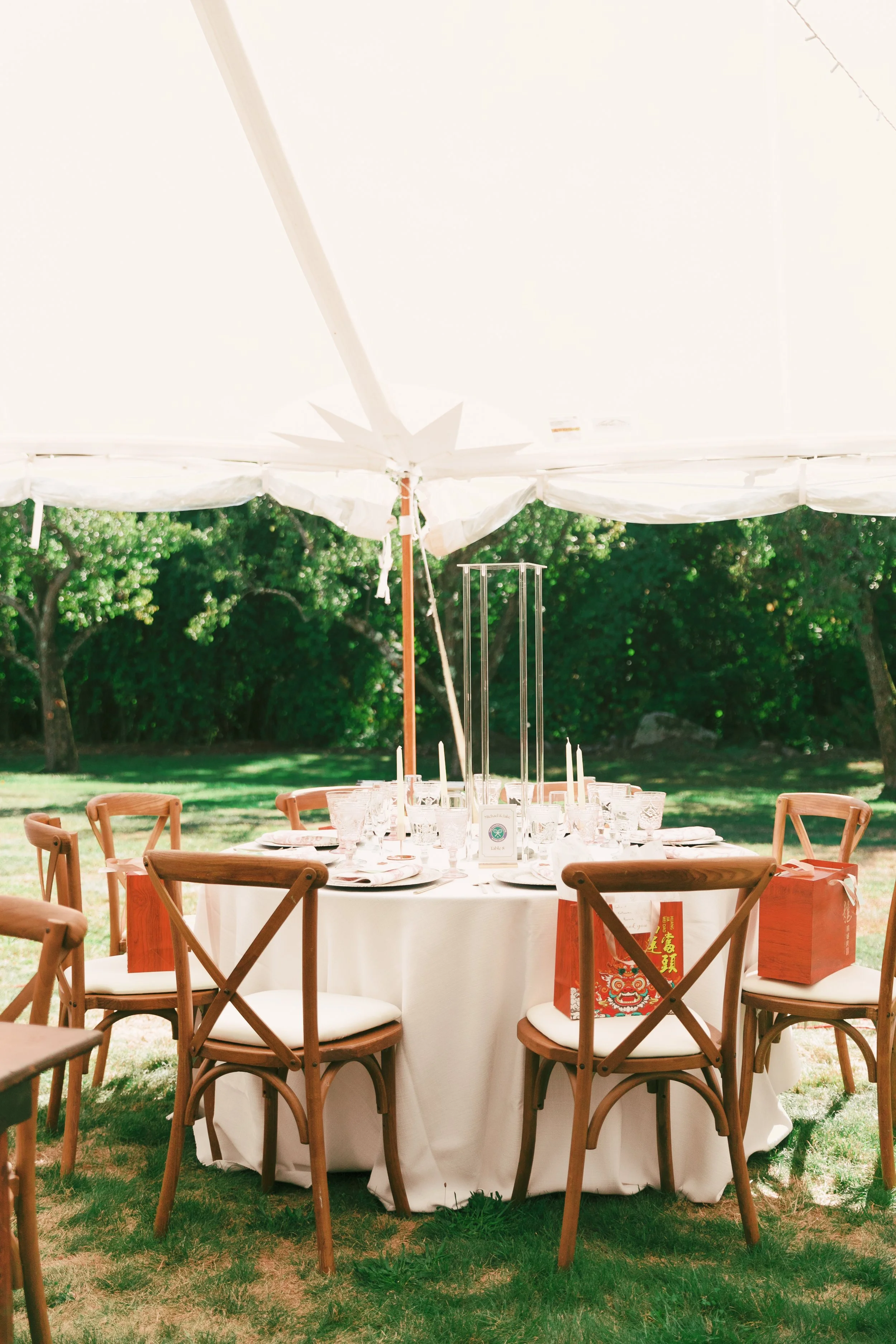 Outdoor event setup with a round table draped in white cloth under a white canopy, surrounded by wooden chairs, set with glassware, plates, candles, and orange gift bags, with a green grassy background and trees.