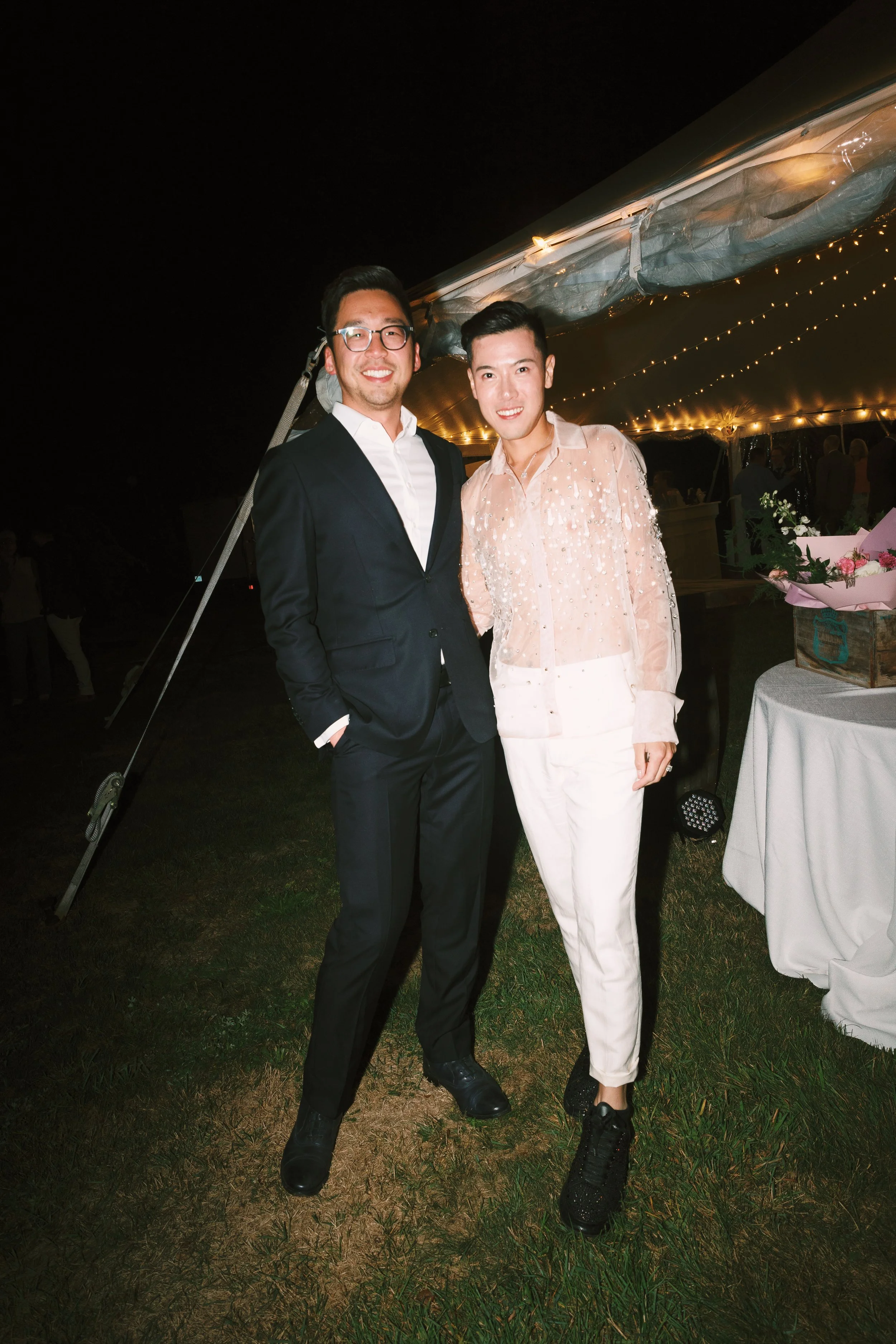 Two men dressed in formal attire standing outdoors at night, smiling, with a decorated tent and tables in the background.