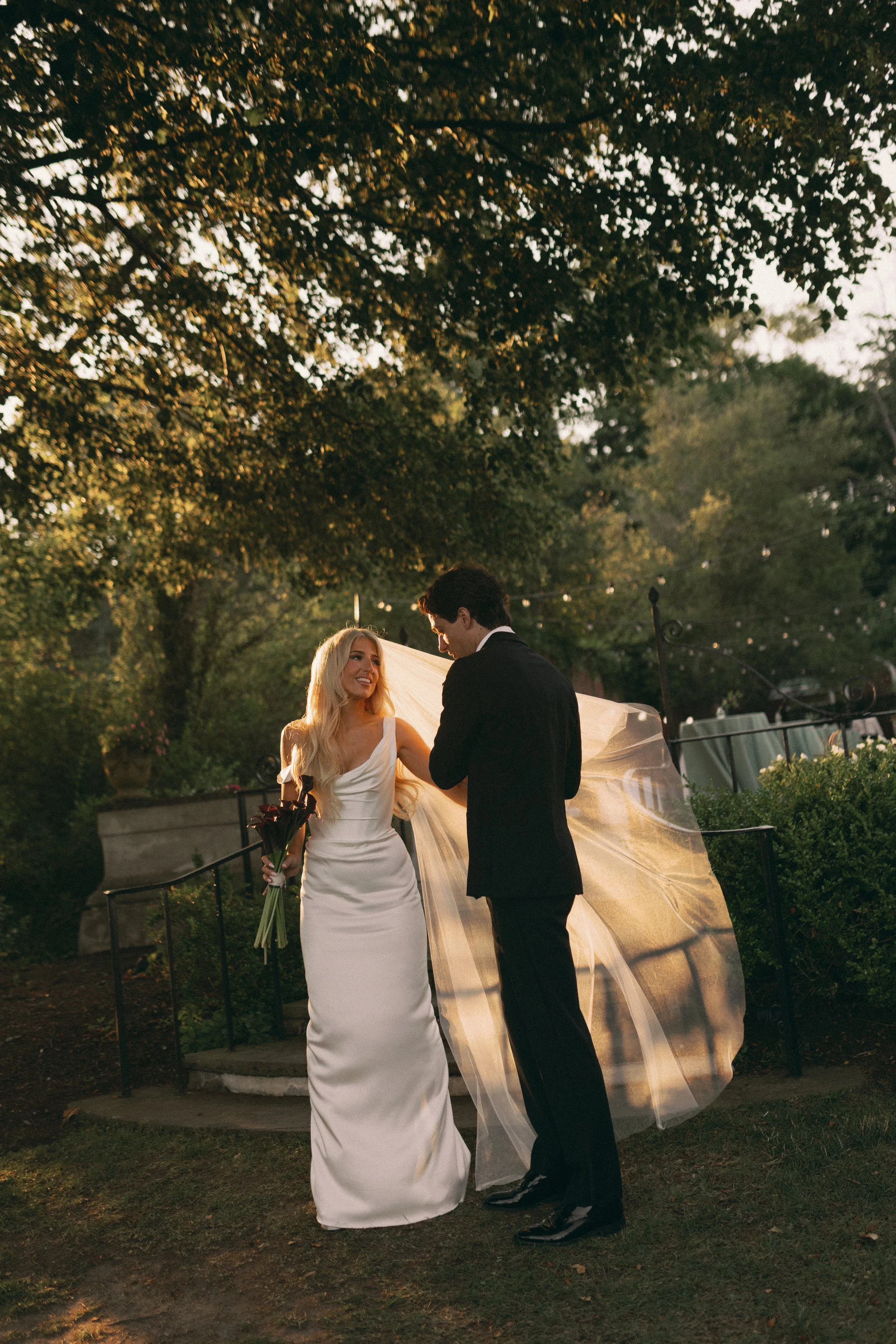 A newlywed couple, a woman in a white wedding dress holding a bouquet, and a man in a black tuxedo, sharing a moment outdoors under a large tree at sunset.