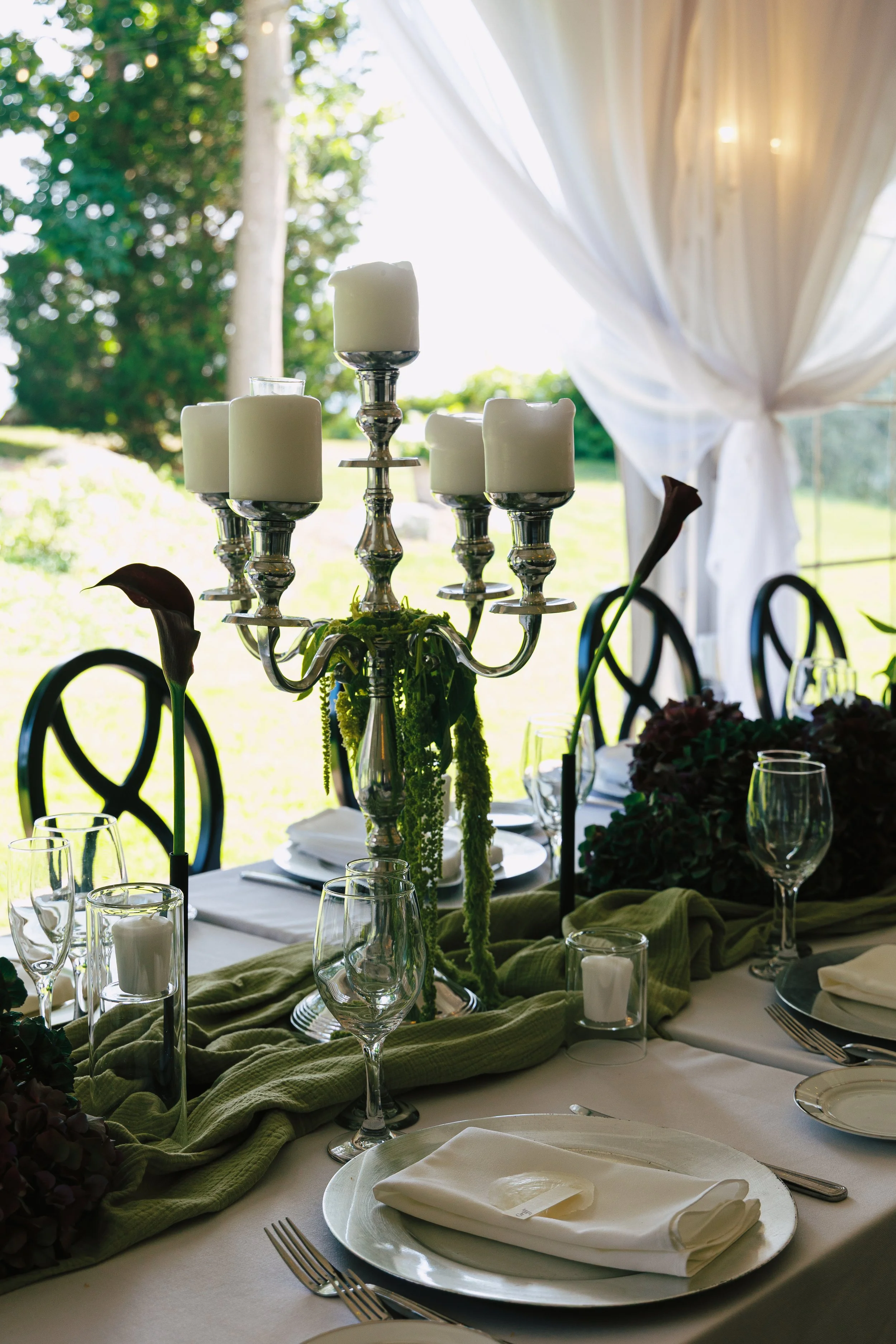 Elegant outdoor table setting with white plates, napkins, wine glasses, and candles, centered around a silver candelabrum with candles, on a table with greenery and a backdrop of trees and a bright sky.