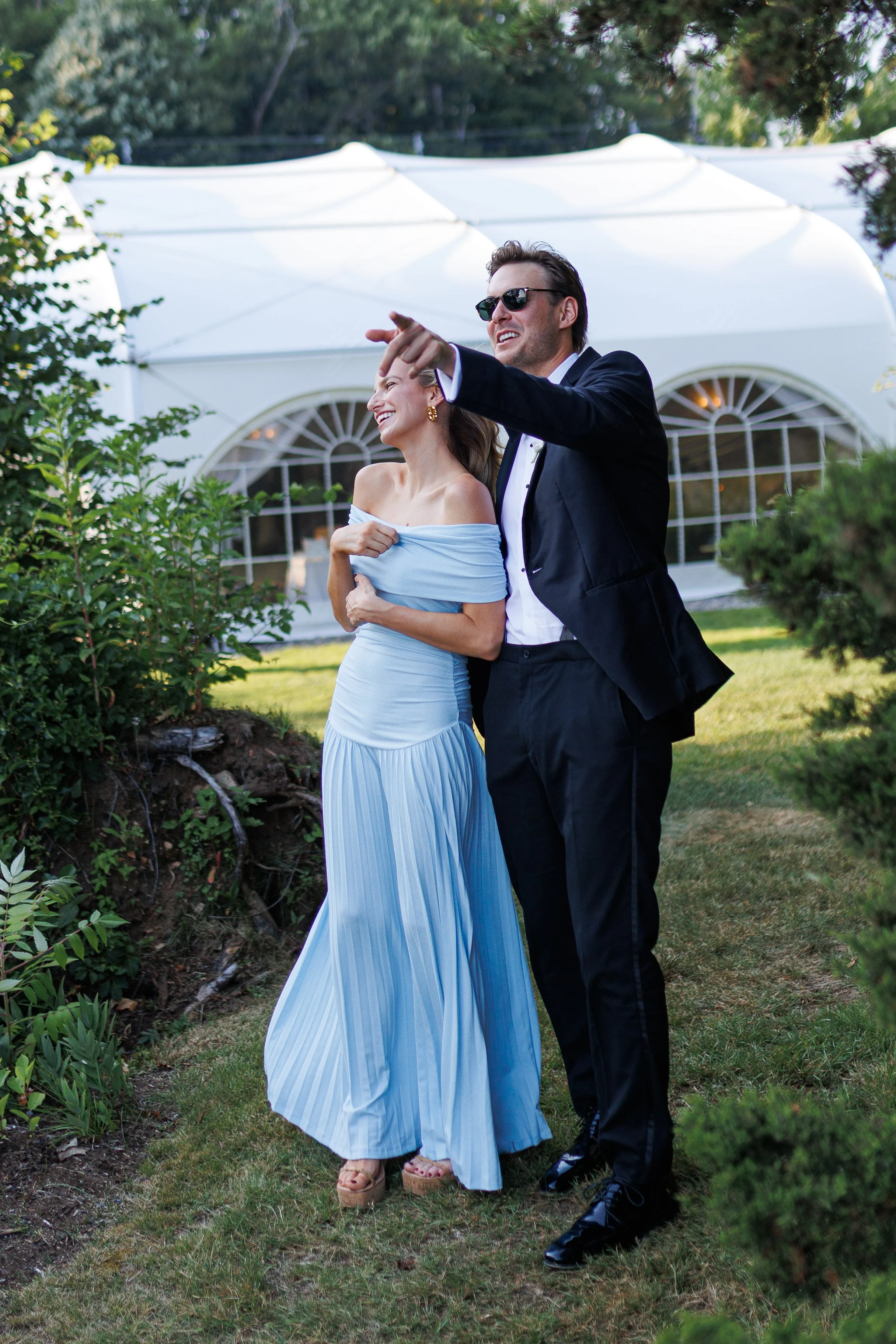 A man and woman dressed formally, standing outdoors in front of a white tent, smiling and pointing at something, enjoying a outdoor event.