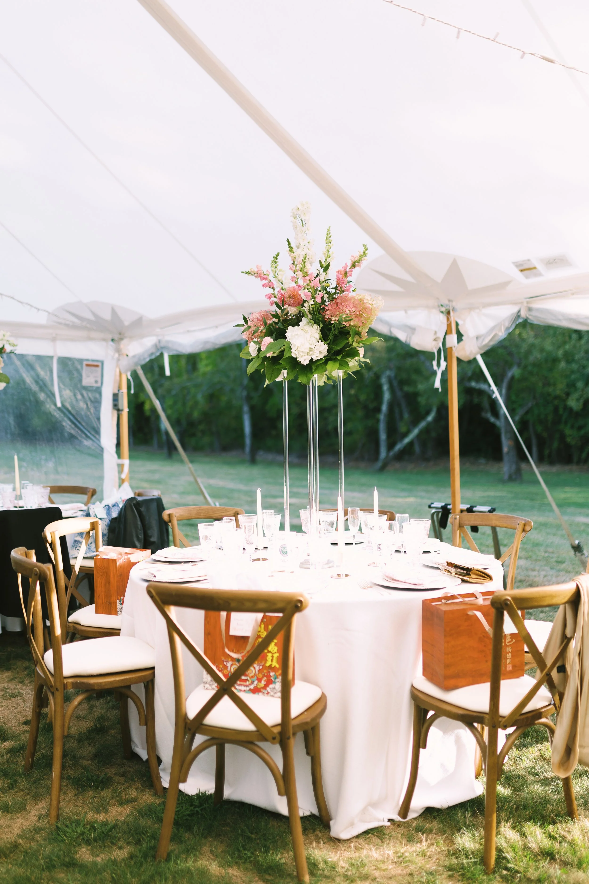 Round outdoor table set for a celebration, decorated with a floral centerpiece, plates, glasses, and candles, under a white canopy tent with wooden chairs around it.