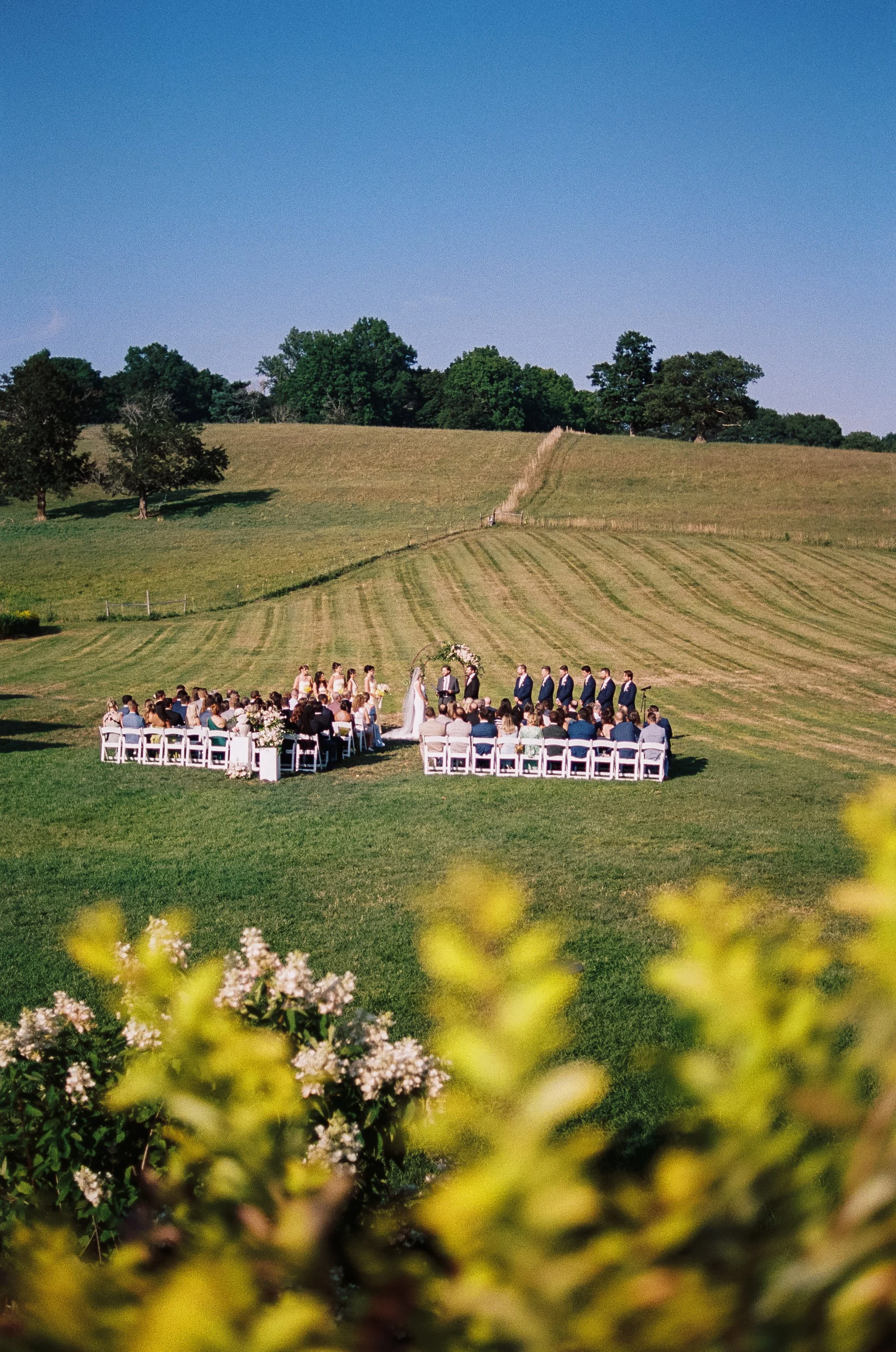 Outdoor wedding ceremony on a grassy field with chairs, officiant, bride, groom, and guests, surrounded by scenic rolling hills and trees under a clear blue sky.