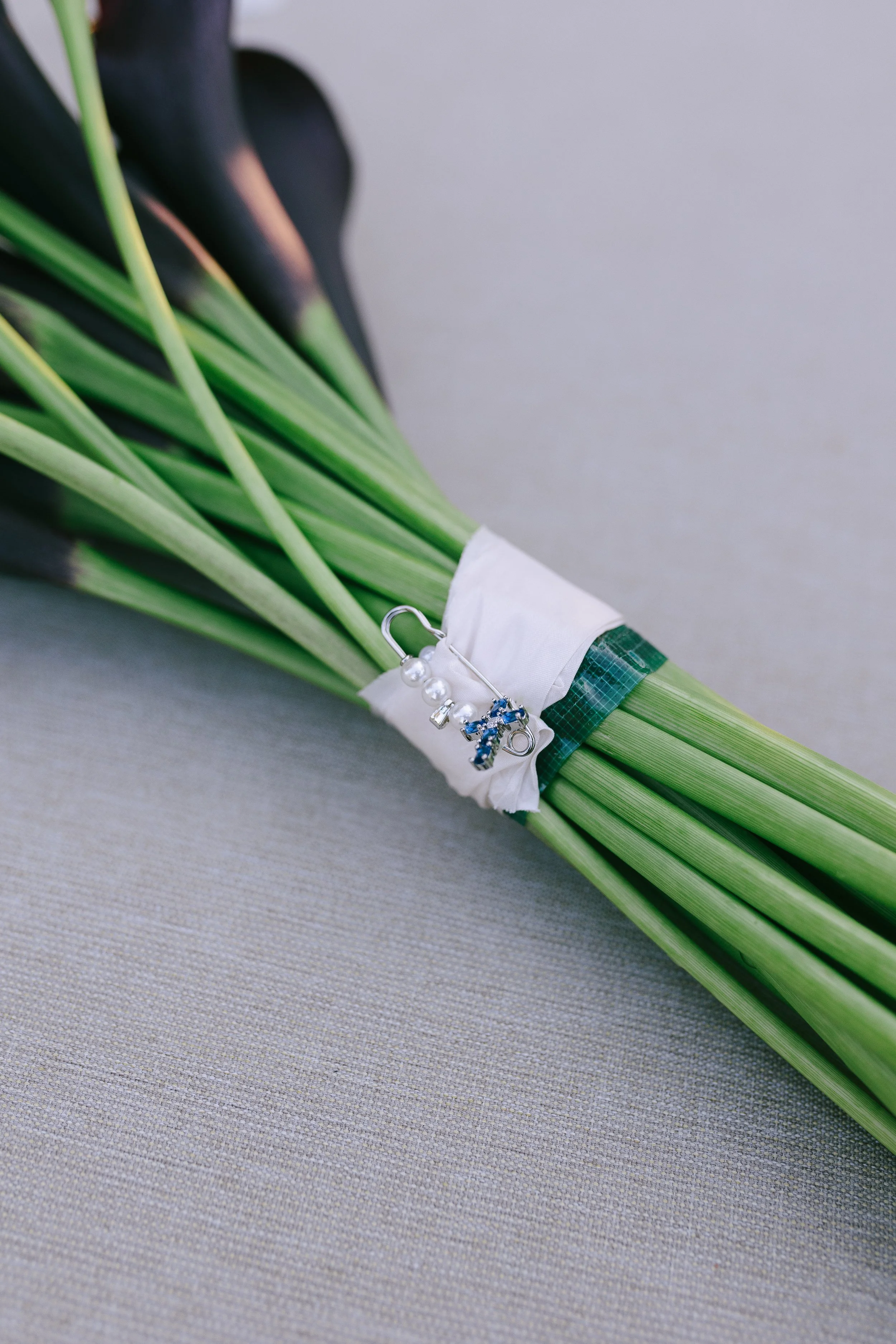 A bouquet of calla lilies wrapped with a white tissue, a green tape, and decorated with a safety pin with pearls and blue stones, lying on a gray fabric surface.