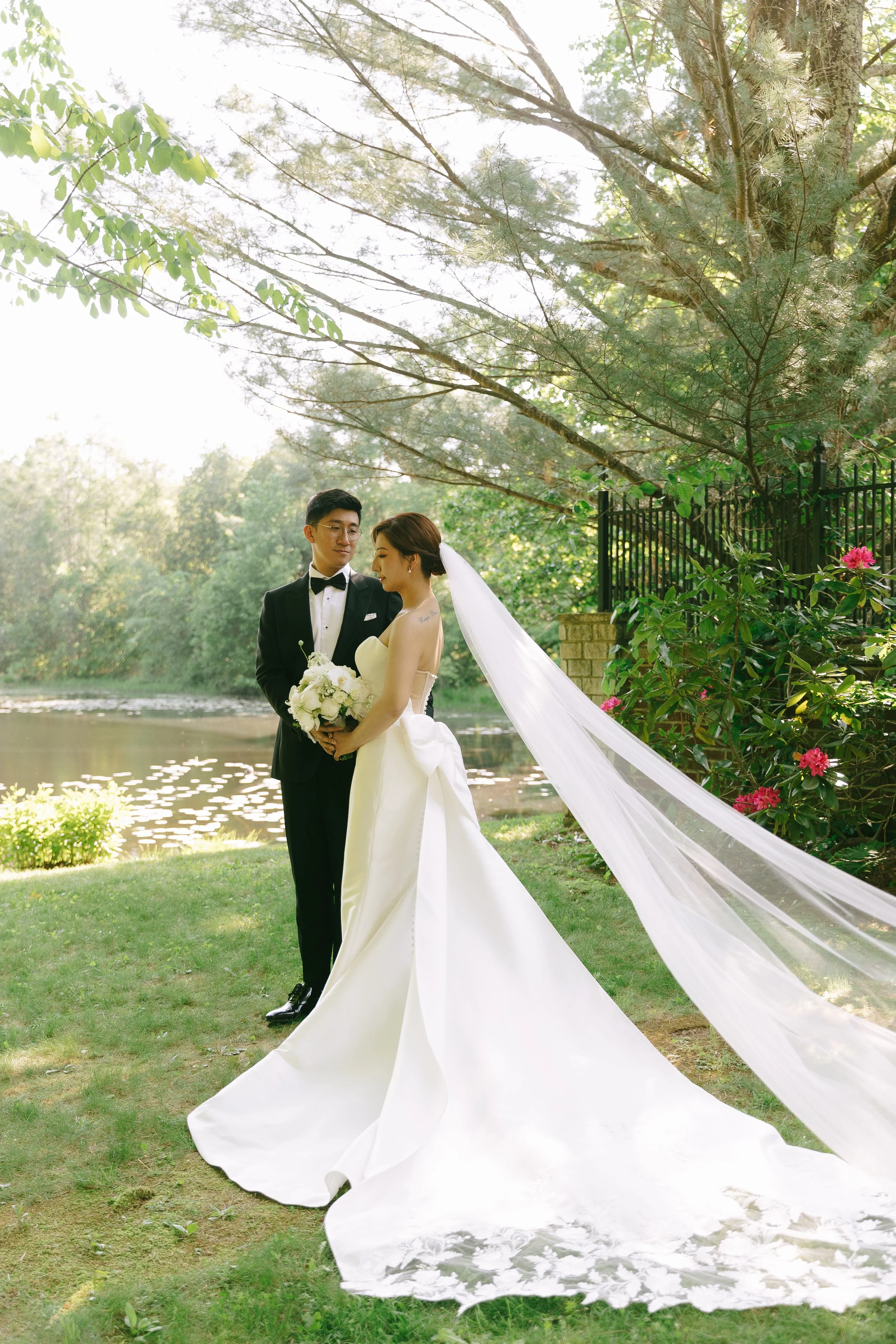 A bride and groom standing by a river in a park, surrounded by trees and flowers, during a wedding photo shoot. The bride is in a white wedding gown with a long veil, holding a bouquet. The groom is in a black tuxedo with a bow tie.