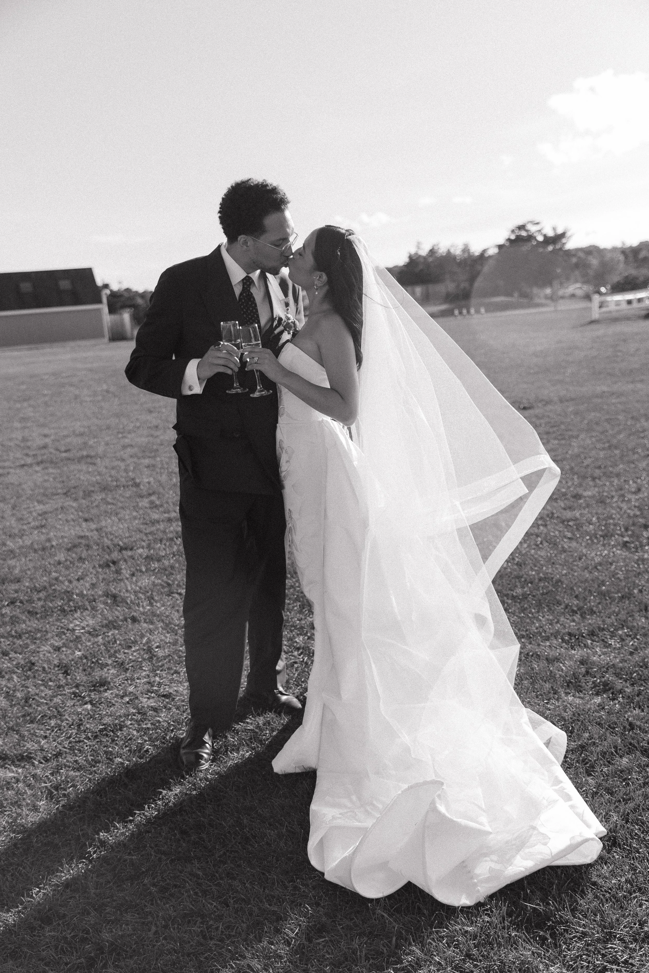 A black and white photo of a bride and groom kissing outdoors, holding champagne glasses on their wedding day.