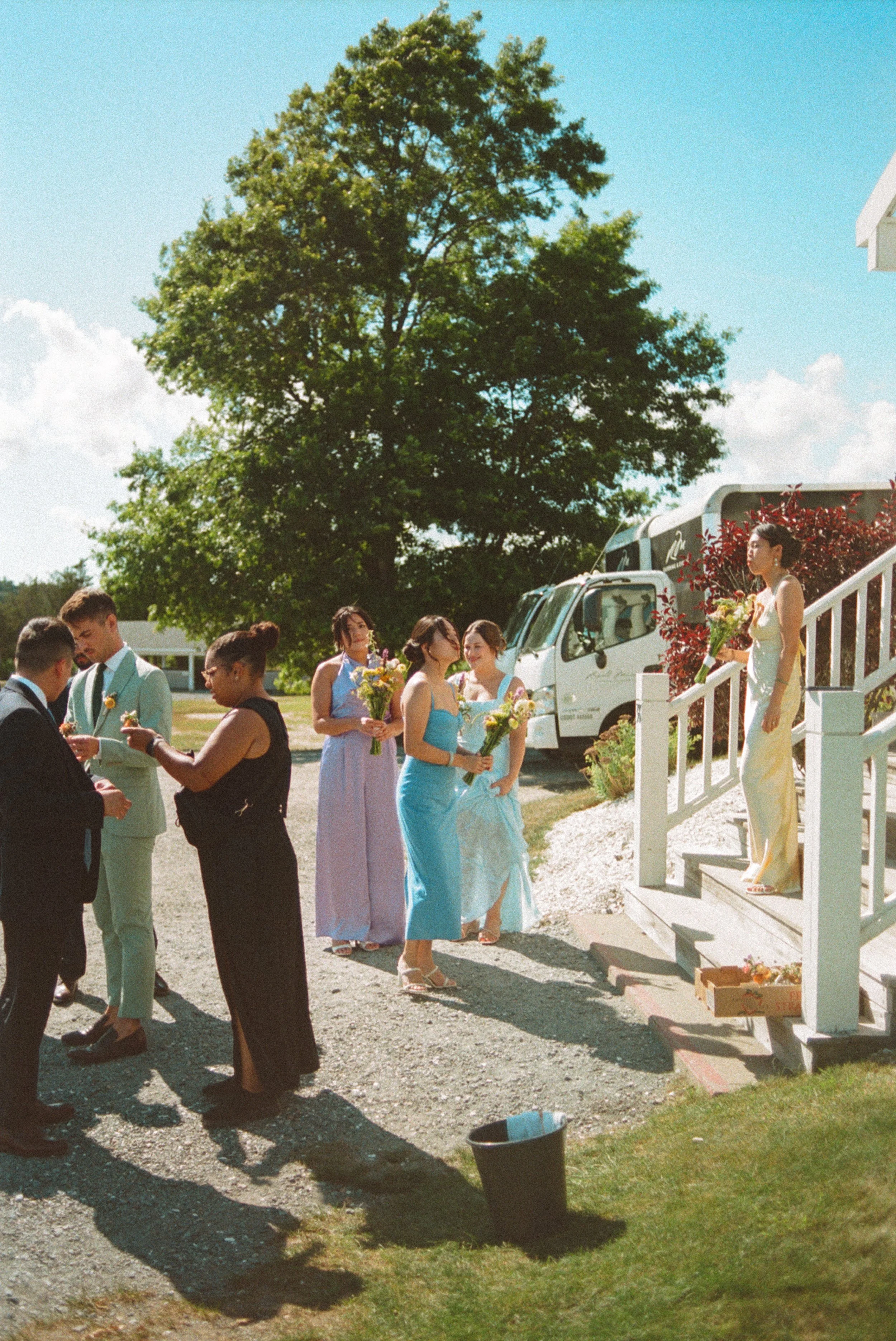 Group of people outdoors near a white staircase with a woman holding flowers standing on the stairs, others holding flowers and glasses, some dressed in formal attire, with a large green tree and a white bus in the background on a sunny day.