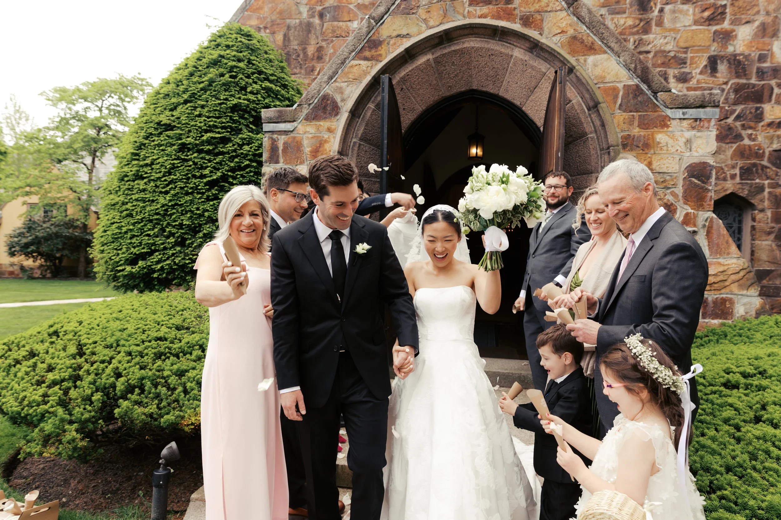A happy wedding celebration with the bride and groom exiting a church, surrounded by family and friends. The bride is holding a large bouquet of white flowers, and the groom is in a black suit. Everyone is smiling and some are holding confetti, with 