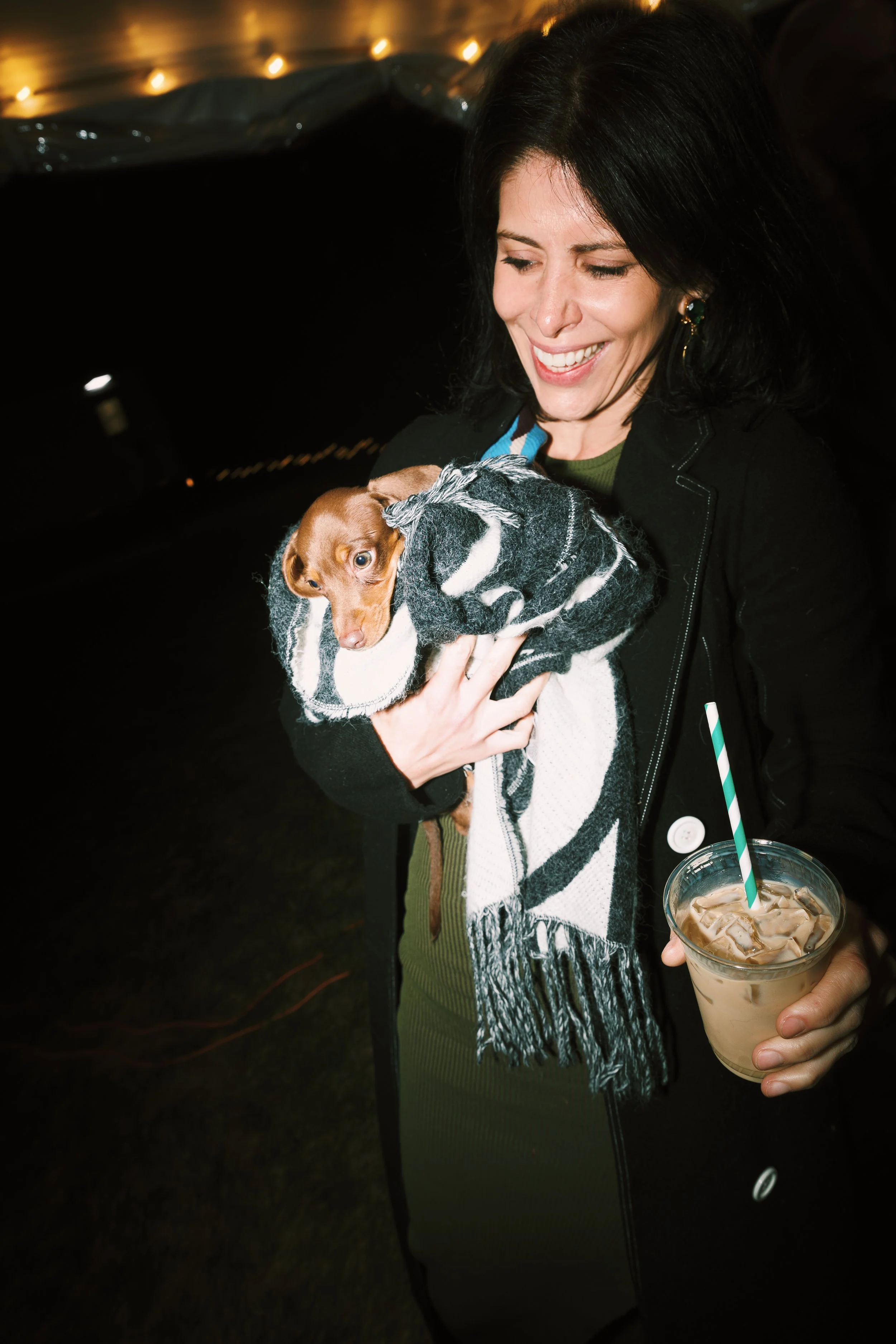 A woman holding a small dog wrapped in a black and white scarf, smiling. She is holding an iced coffee with a straw and wearing a black coat.