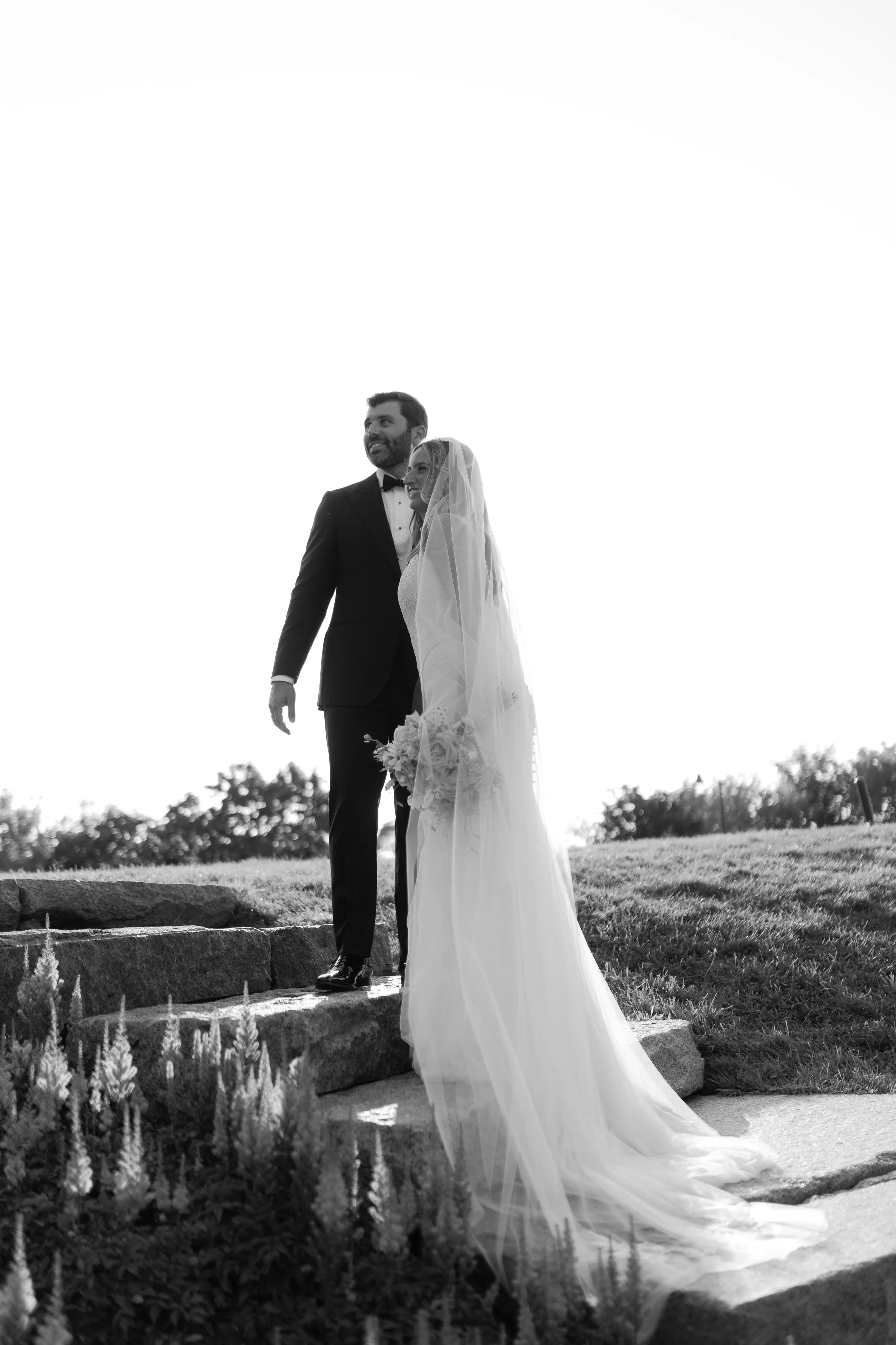 Black and white photo of a bride and groom standing on stone steps outdoors, smiling, with the bride holding a bouquet and wearing a veil, and the groom in a tuxedo, scenic background, bright sky.
