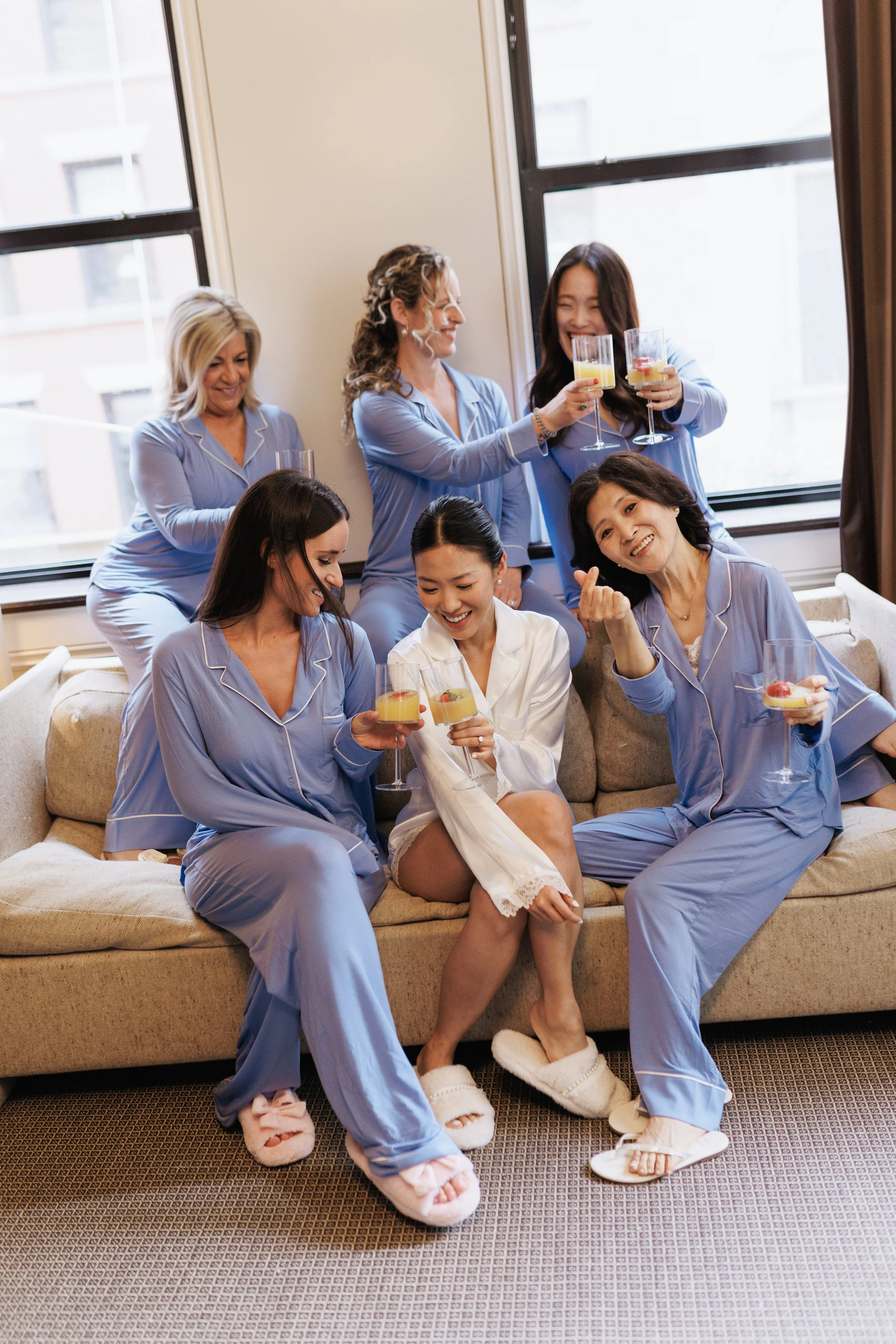 A group of seven women in pajamas celebrating with drinks in a bright room with large windows.