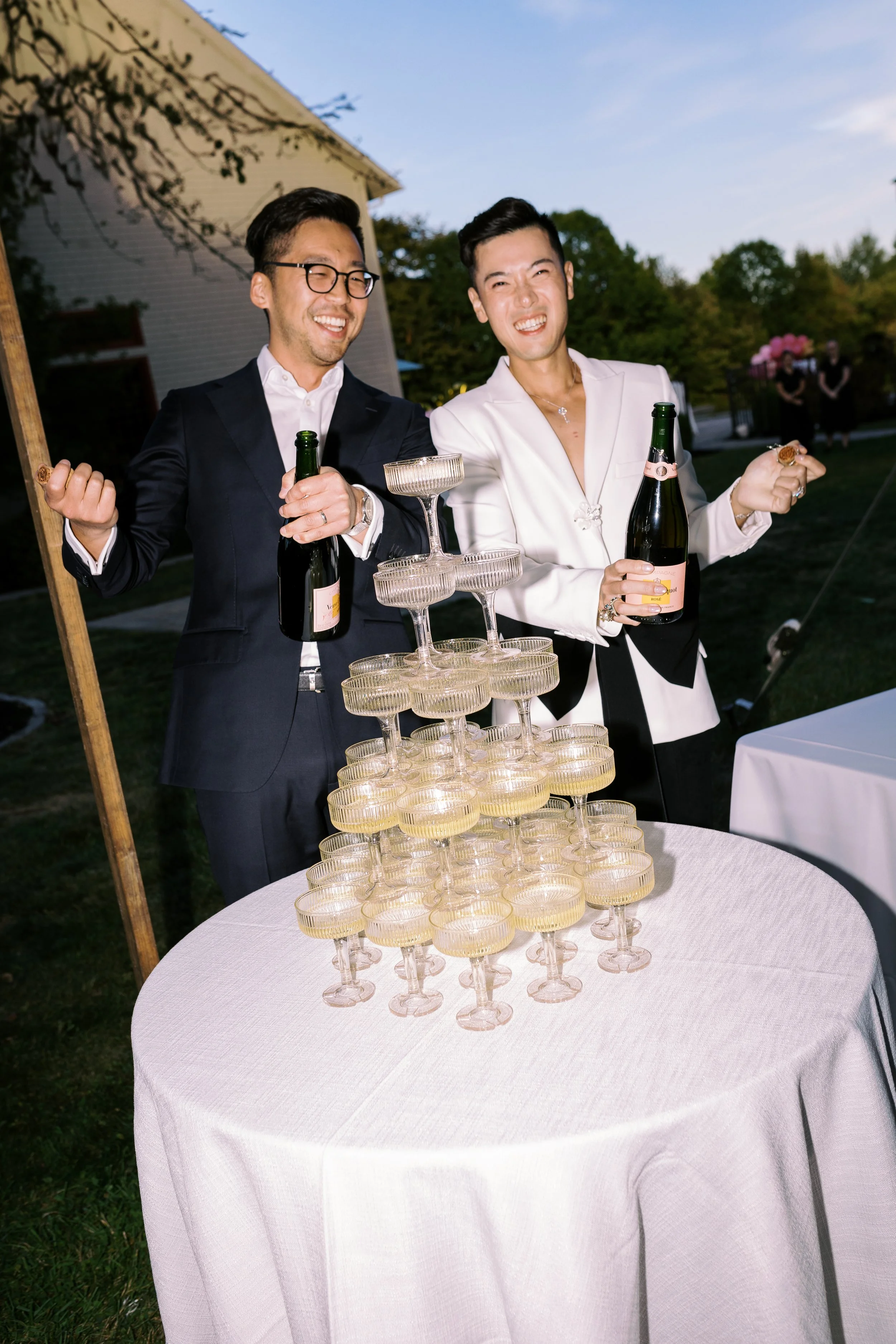 Two men celebrating at an outdoor event, pouring champagne into a pyramid of glasses on a white table.