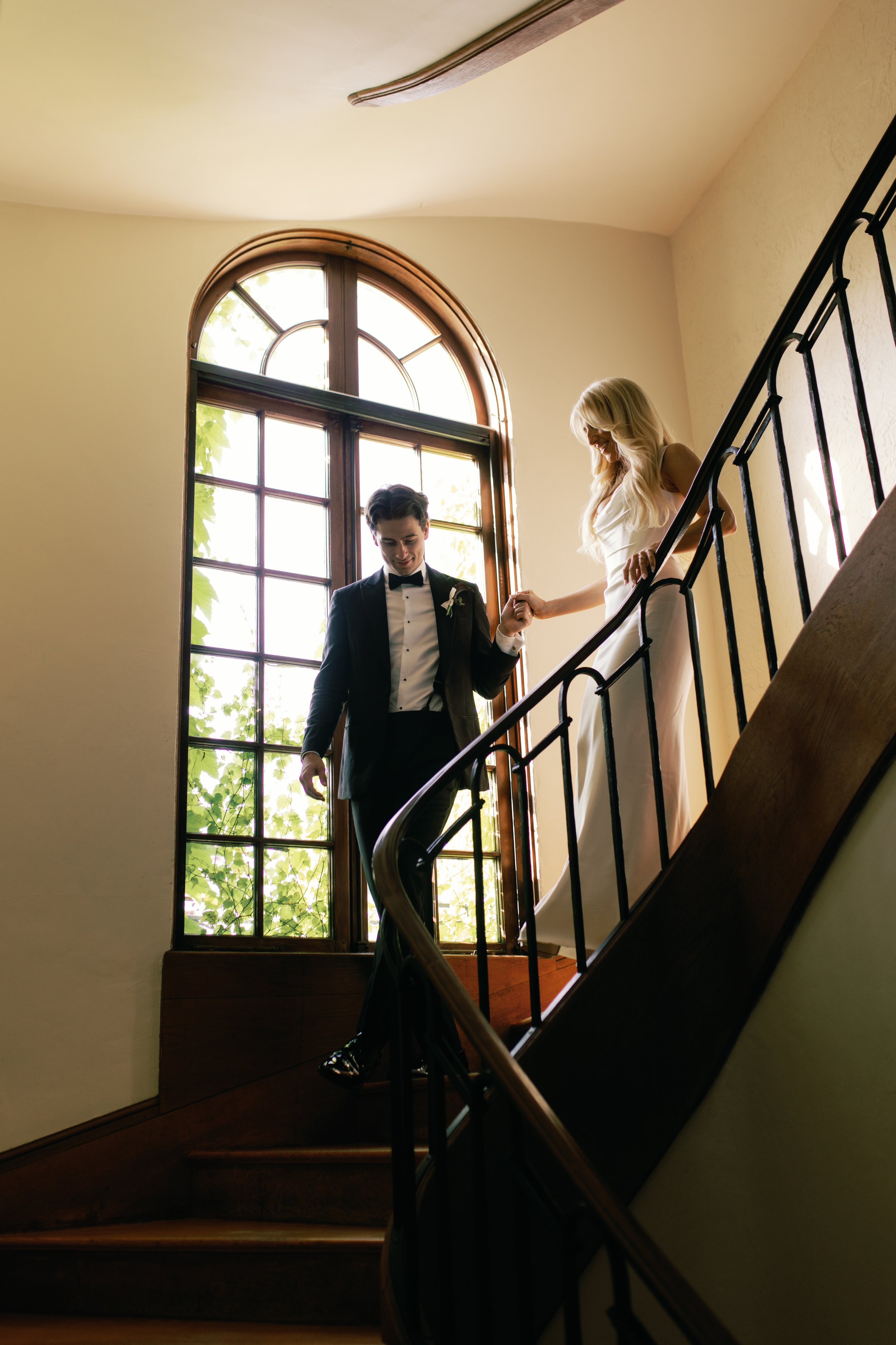 A bride and groom descending a staircase holding hands, near a large arched window with sunlight shining through.