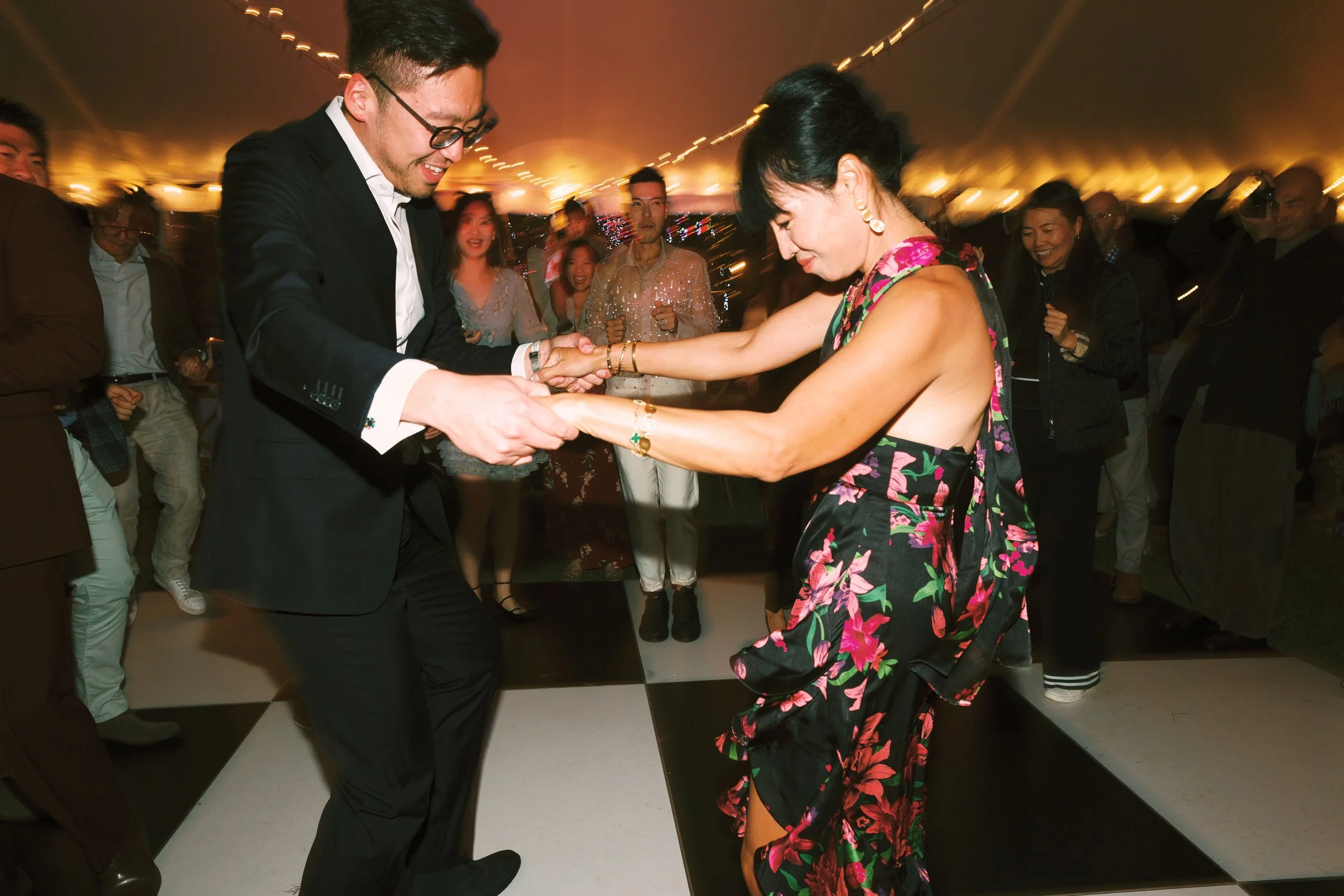 A man and woman are dancing together at a lively party, holding hands and smiling, with several people watching and celebrating in the background under warm lighting.