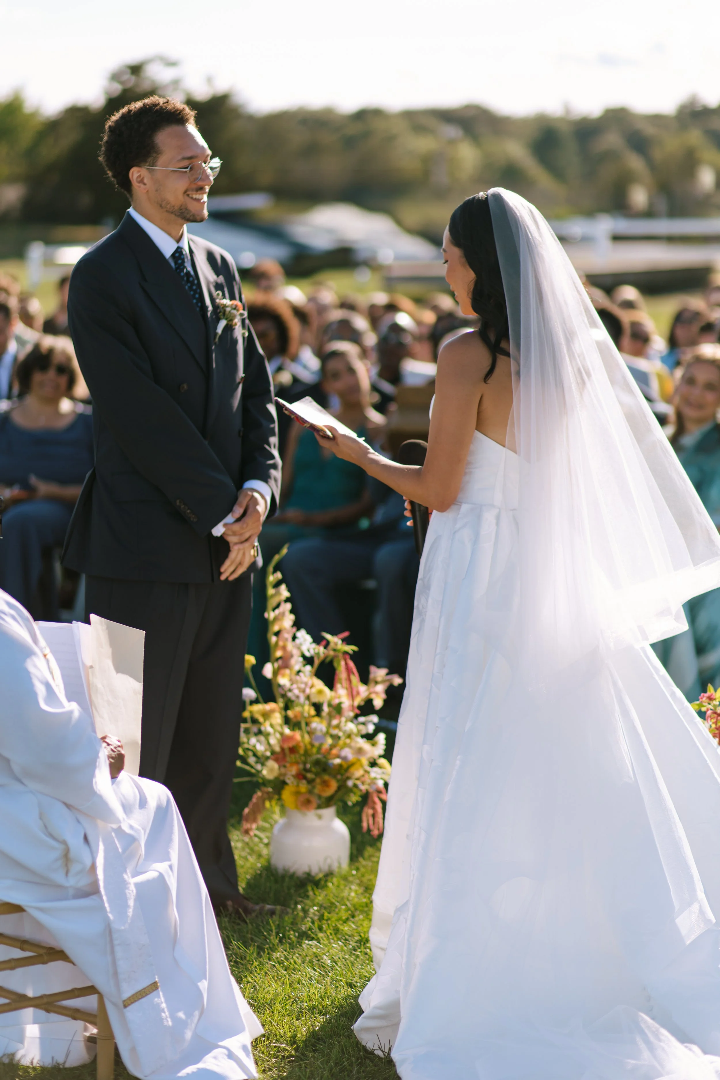 A bride and groom exchanging vows during an outdoor wedding ceremony; the bride is wearing a white wedding gown and veil, and the groom is in a dark suit with glasses, with guests seated behind them.
