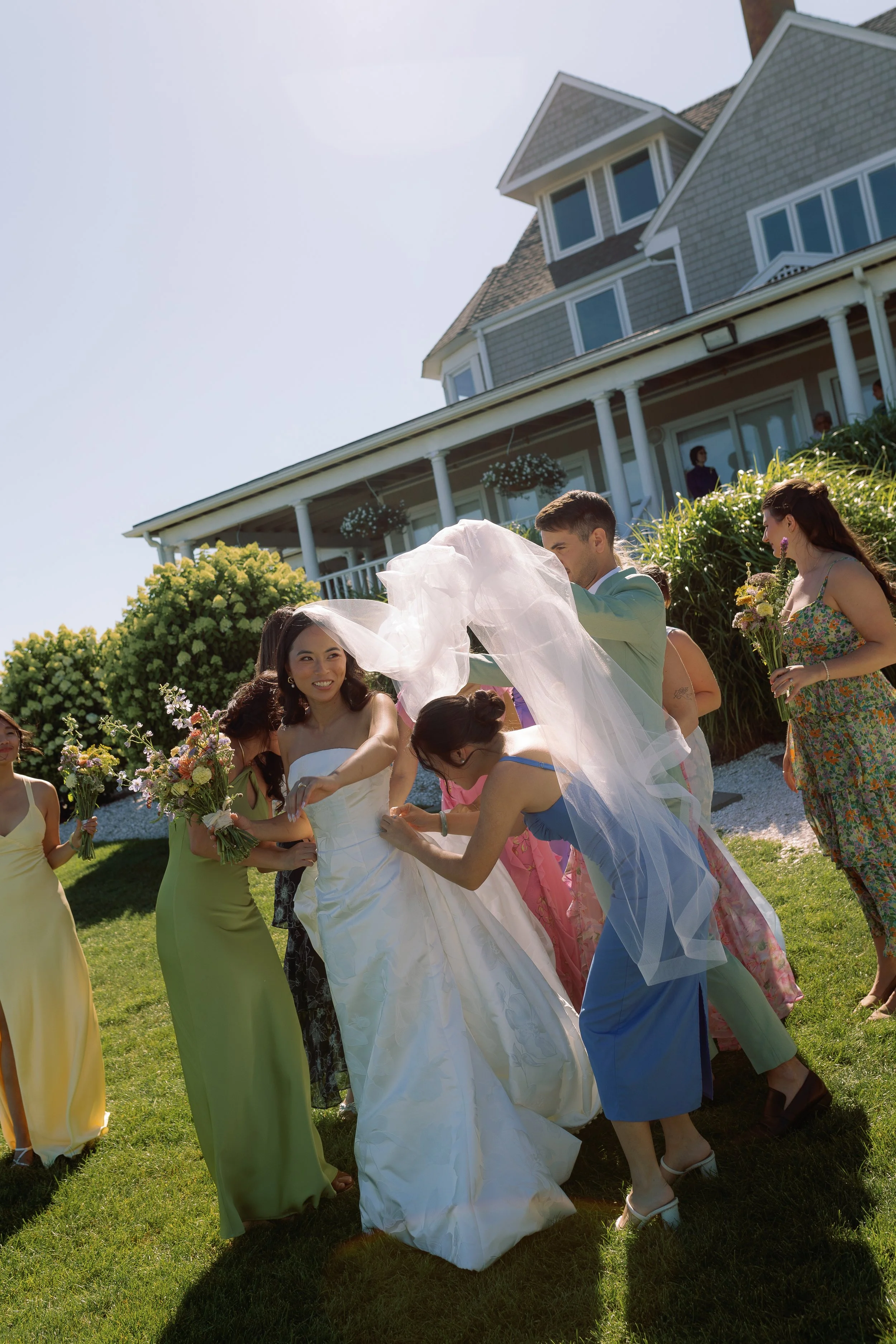 A bride in a white wedding dress getting ready for a wedding with the help of bridesmaids and friends outside a large house on a sunny day.
