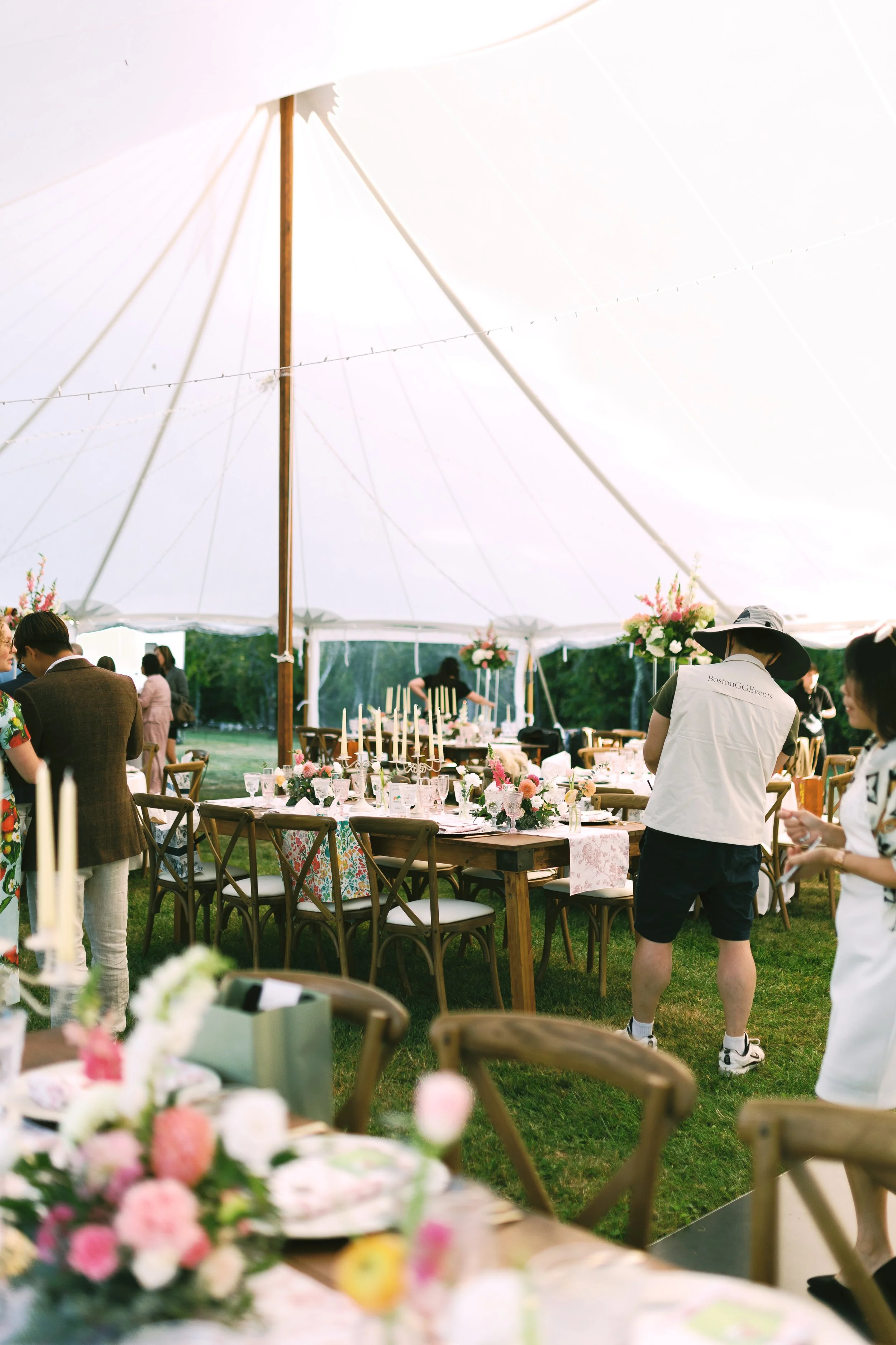 A large outdoor wedding reception under a white tent with decorated tables, floral arrangements, and candles, with people preparing and setting up.
