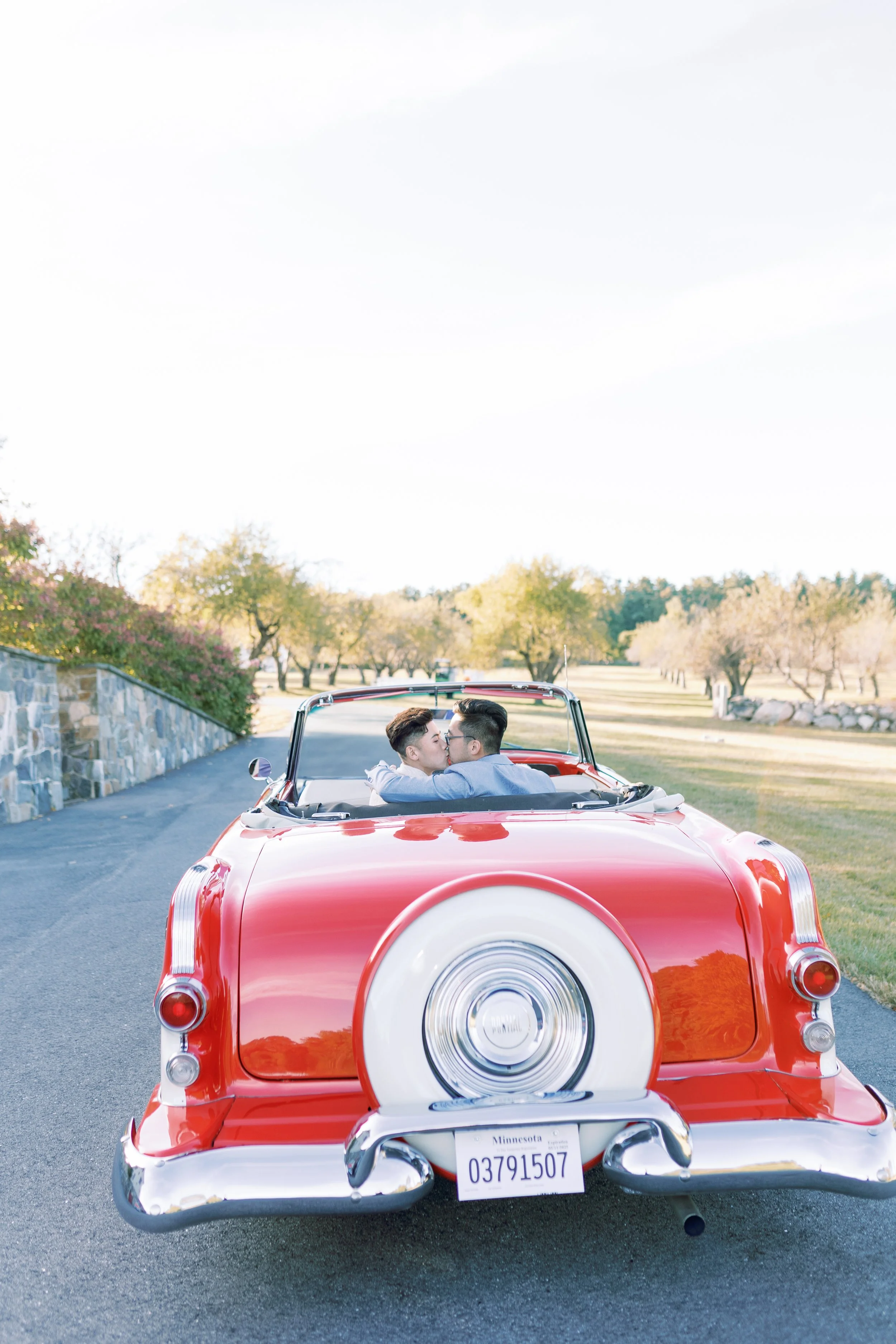 A couple kissing in a vintage red convertible car parked on a paved road with trees and a stone wall in the background.