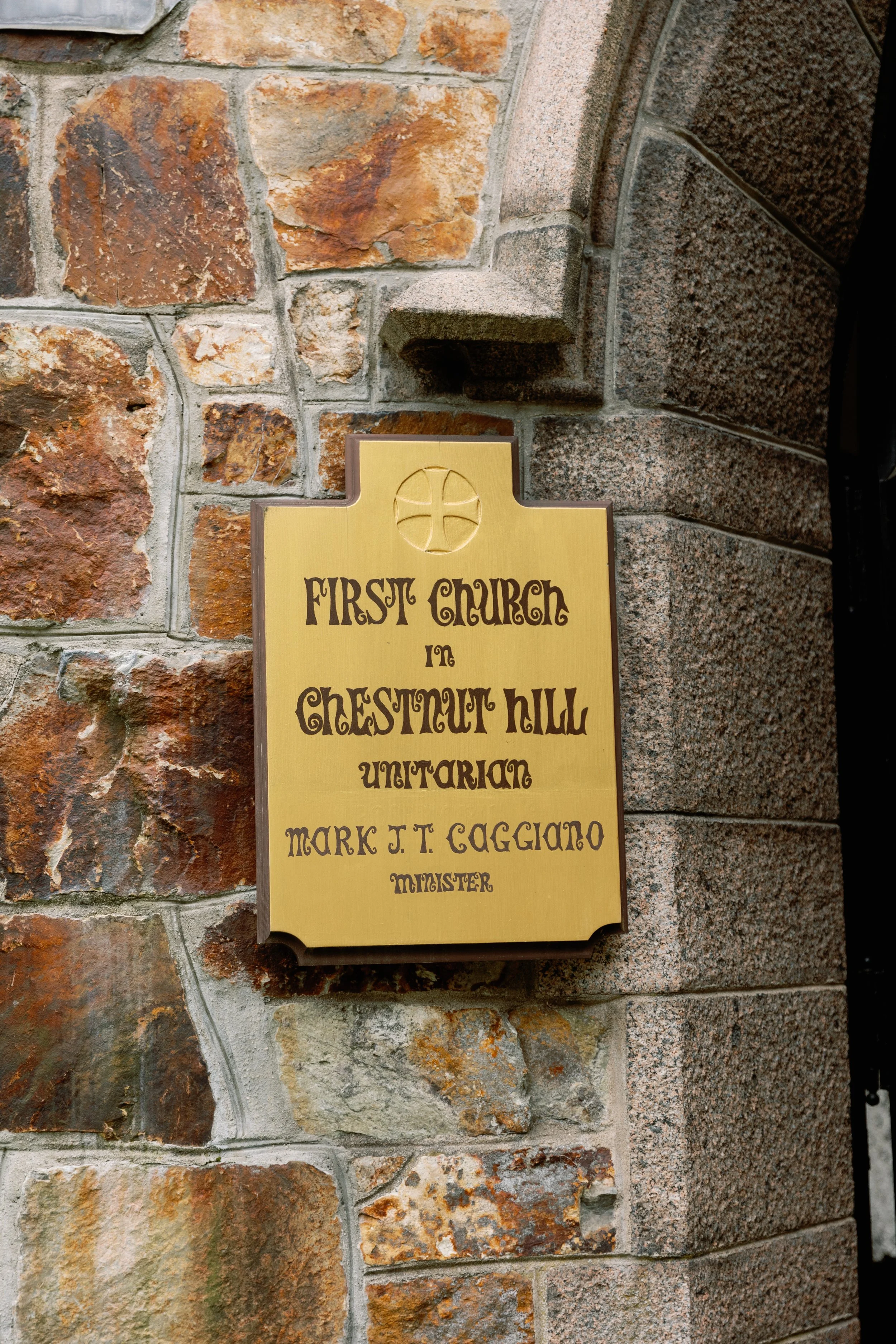 A sign on a brick church wall that reads, 'First Church in Chestnut Hill Unitarian, Mark J. T. Caggiano Minister,' with a cross symbol above the text.