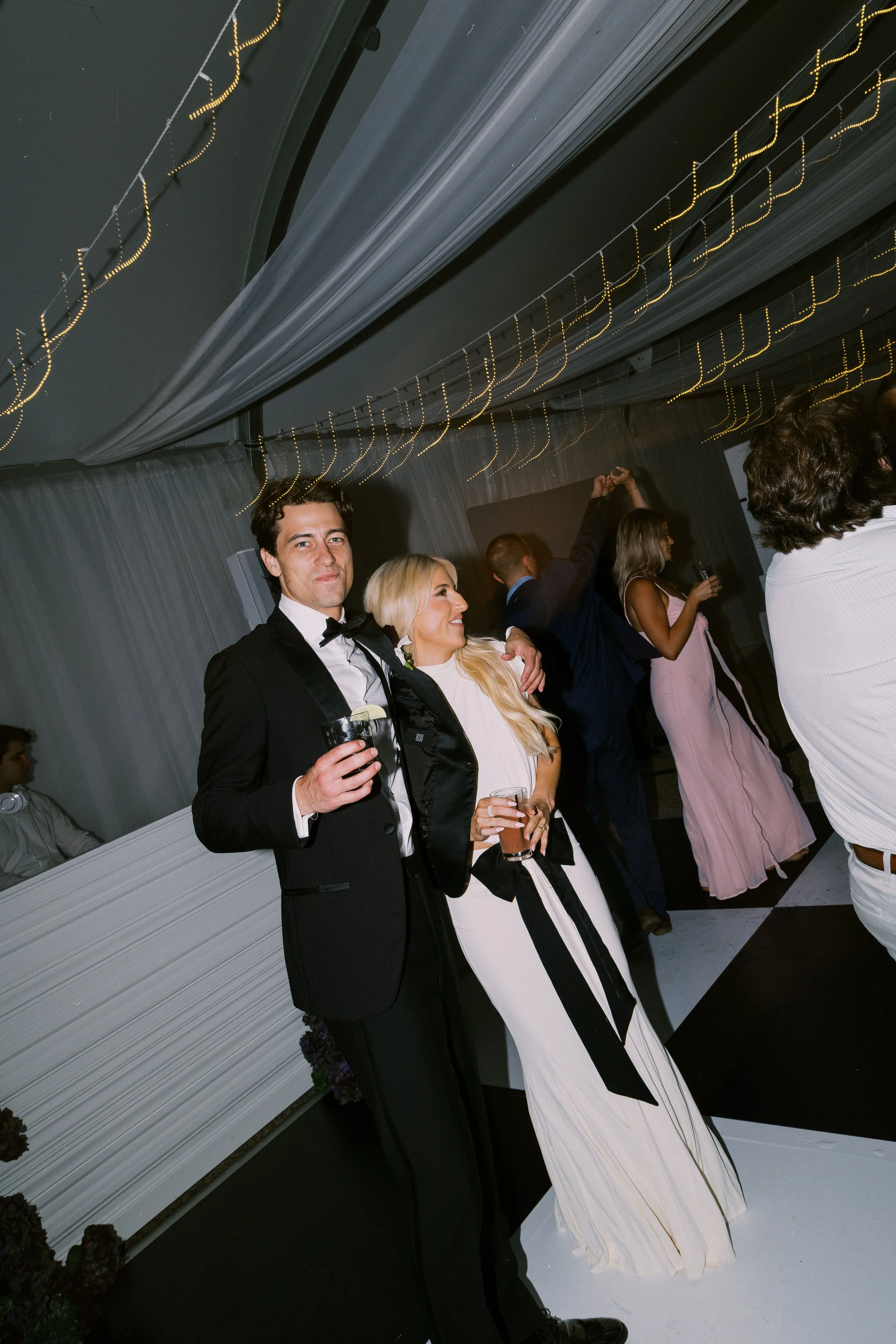 A couple dressed in formal attire, with the man in a tuxedo and the woman in a white dress, enjoying drinks at a party or wedding reception with string lights and other guests dancing in the background.