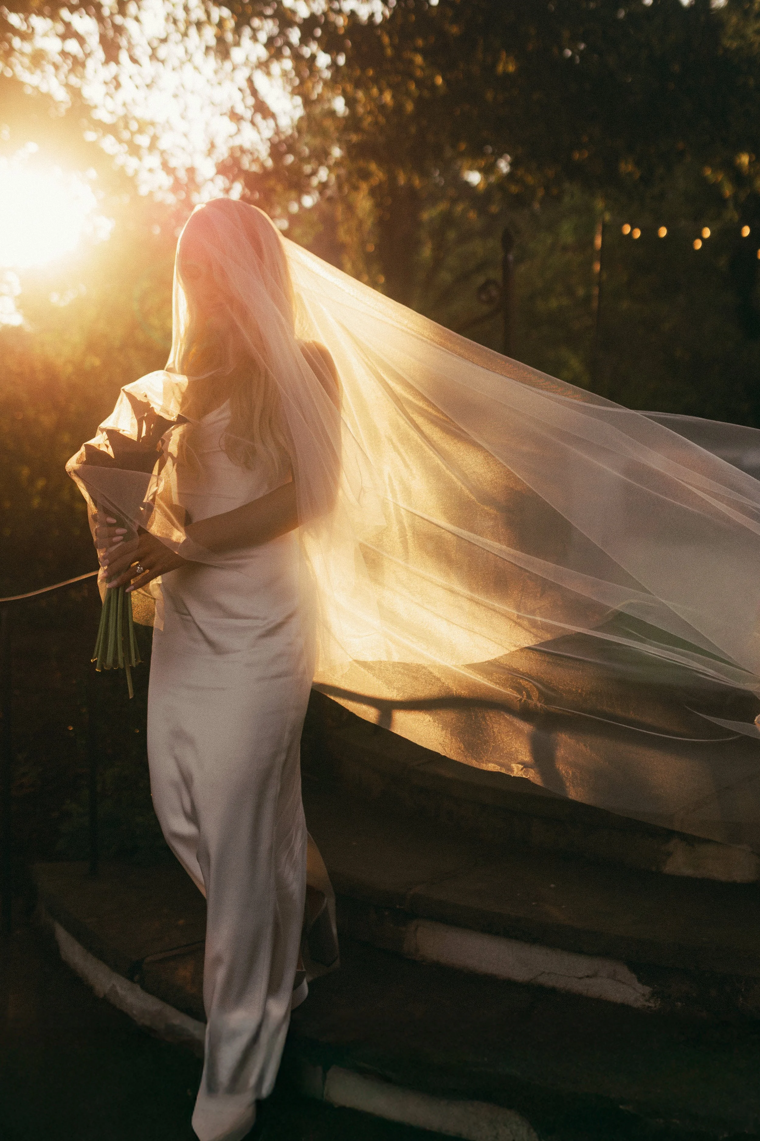 A bride holding a bouquet of flowers during sunset, standing outdoors with trees and string lights in the background, wearing a white wedding dress and veil.