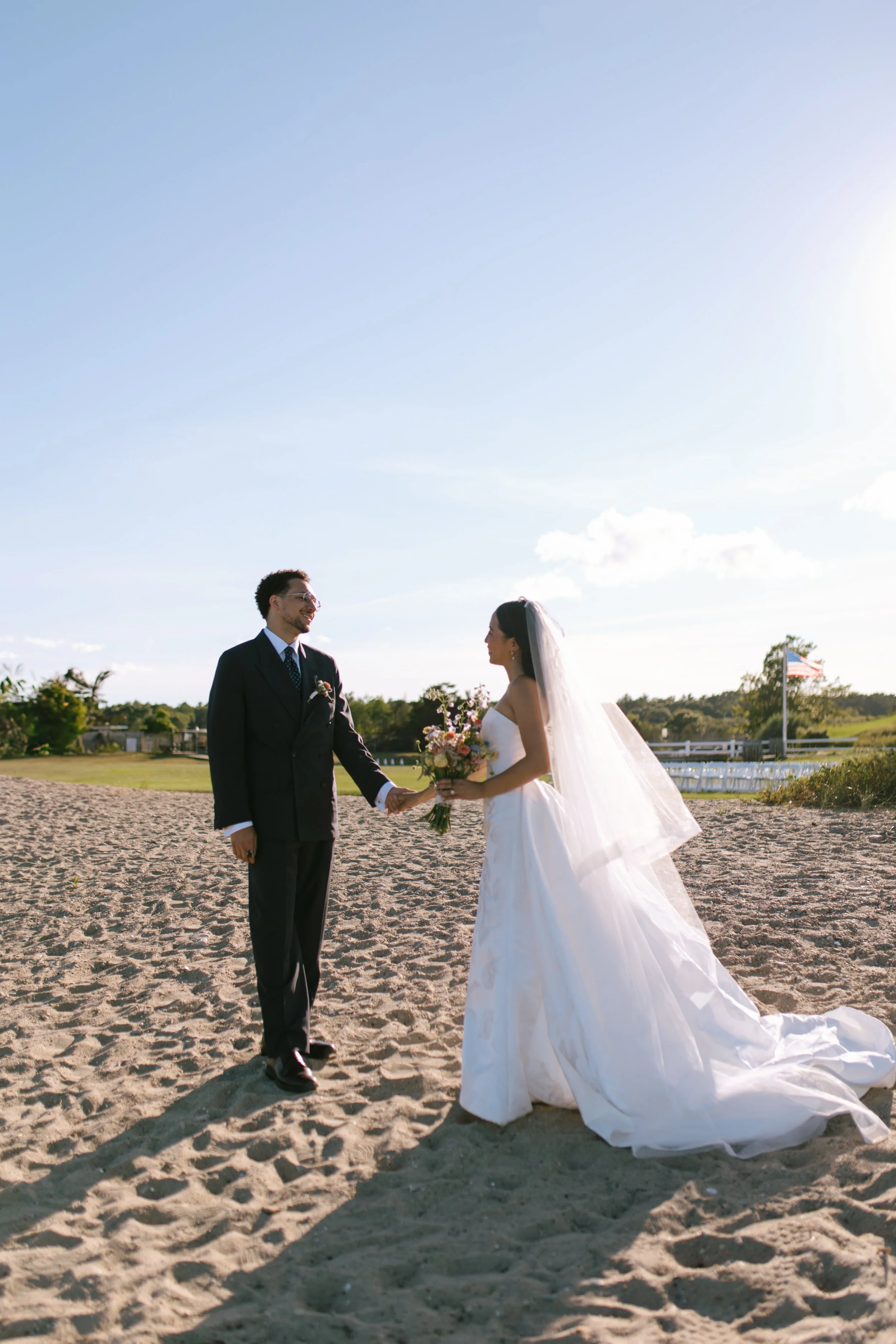 A bride and groom holding hands on a sandy beach during sunset, with the groom in a dark suit and the bride in a strapless white wedding gown with a long train and veil, holding a bouquet of flowers.