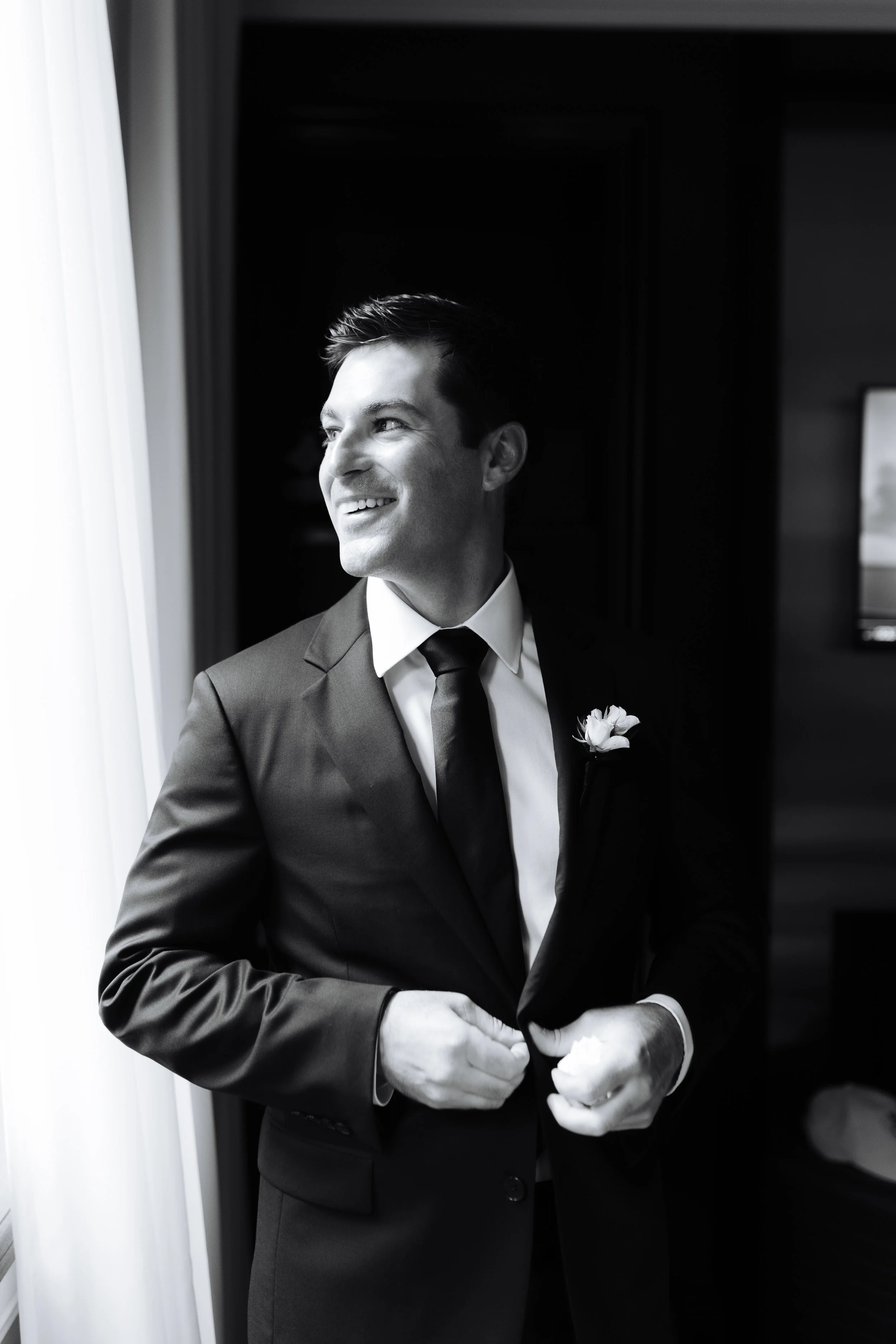 A man in a suit and tie, smiling and standing by a window, adjusting his suit jacket. It's a black-and-white photo.