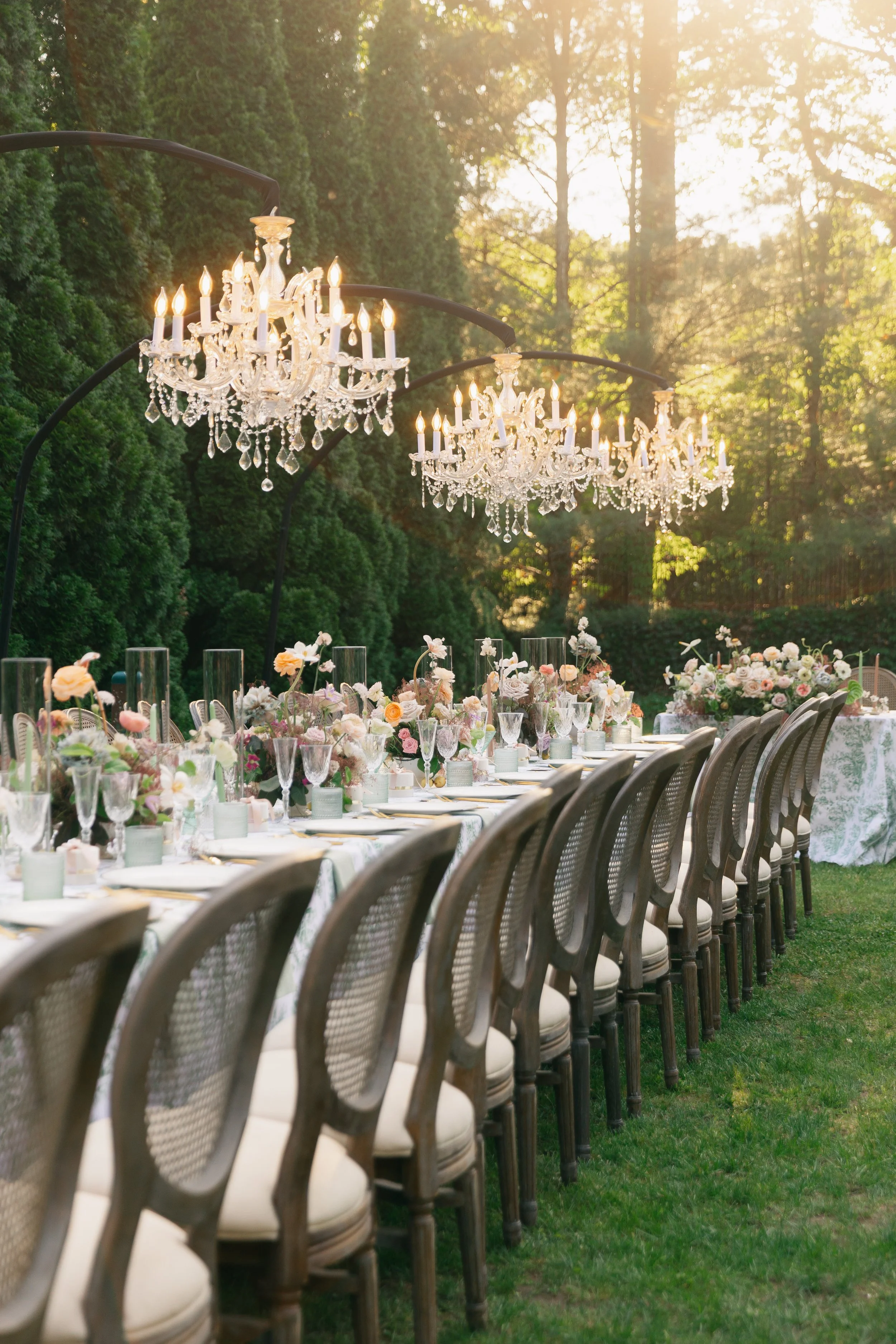 A long outdoor banquet table decorated with floral centerpieces and fine tableware, set beneath two chandeliers in a lush green garden with sunlight filtering through trees.