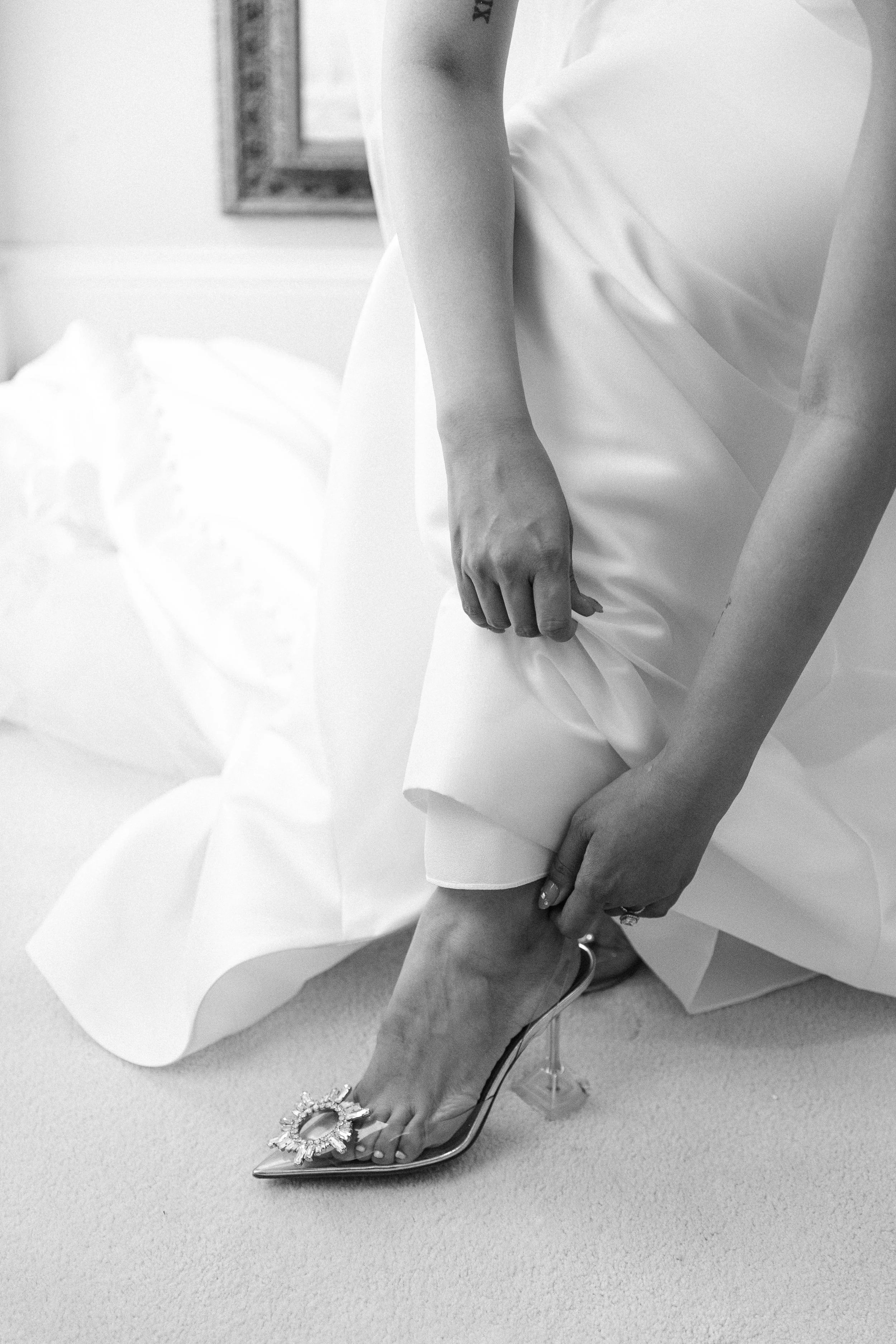 A woman in a wedding dress adjusts her high-heeled shoe with a decorative embellishment.