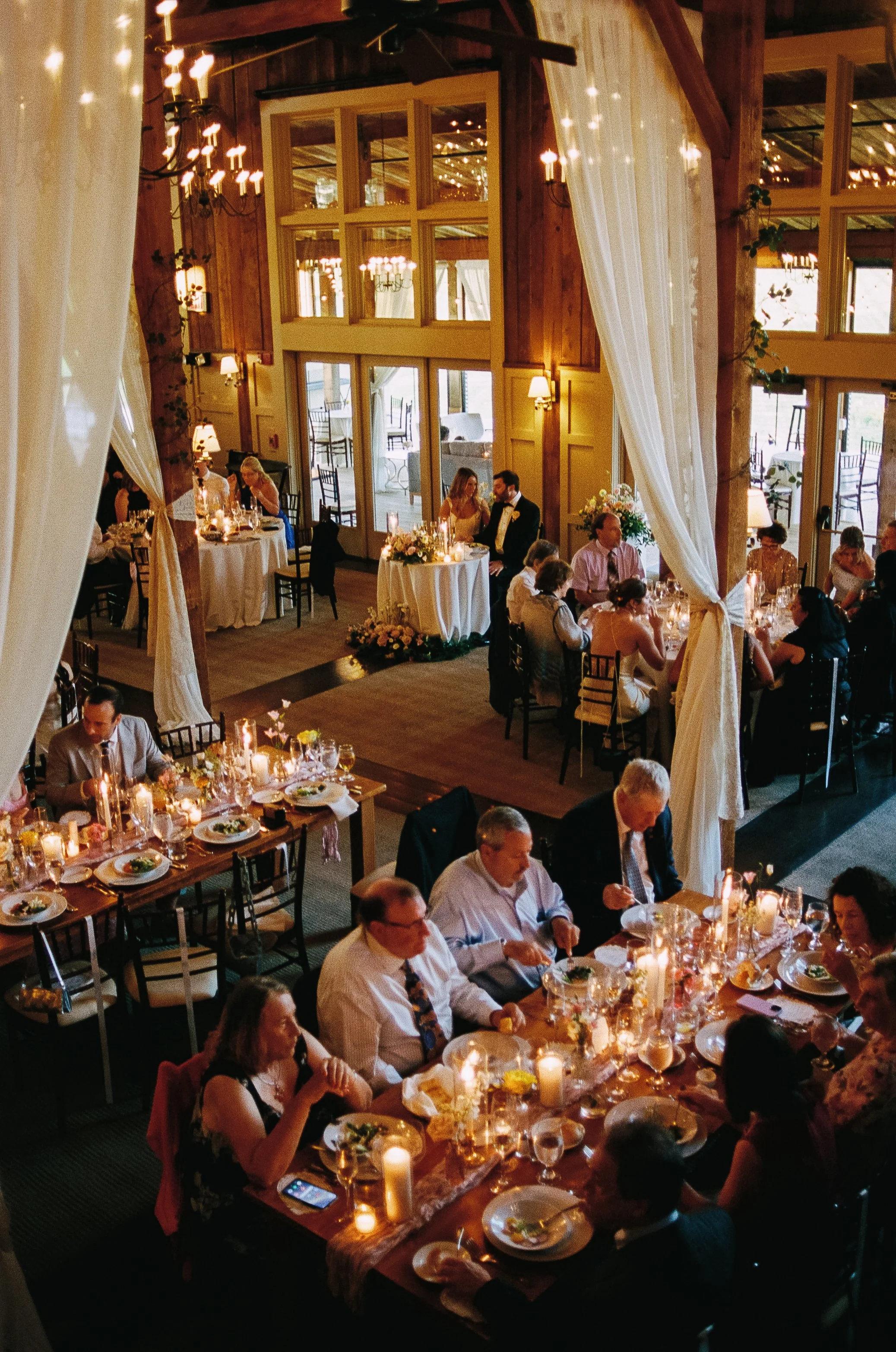 A large indoor wedding reception with guests seated at decorated tables, candles, floral centerpieces, and large windows with flowing curtains in a rustic wooden venue.