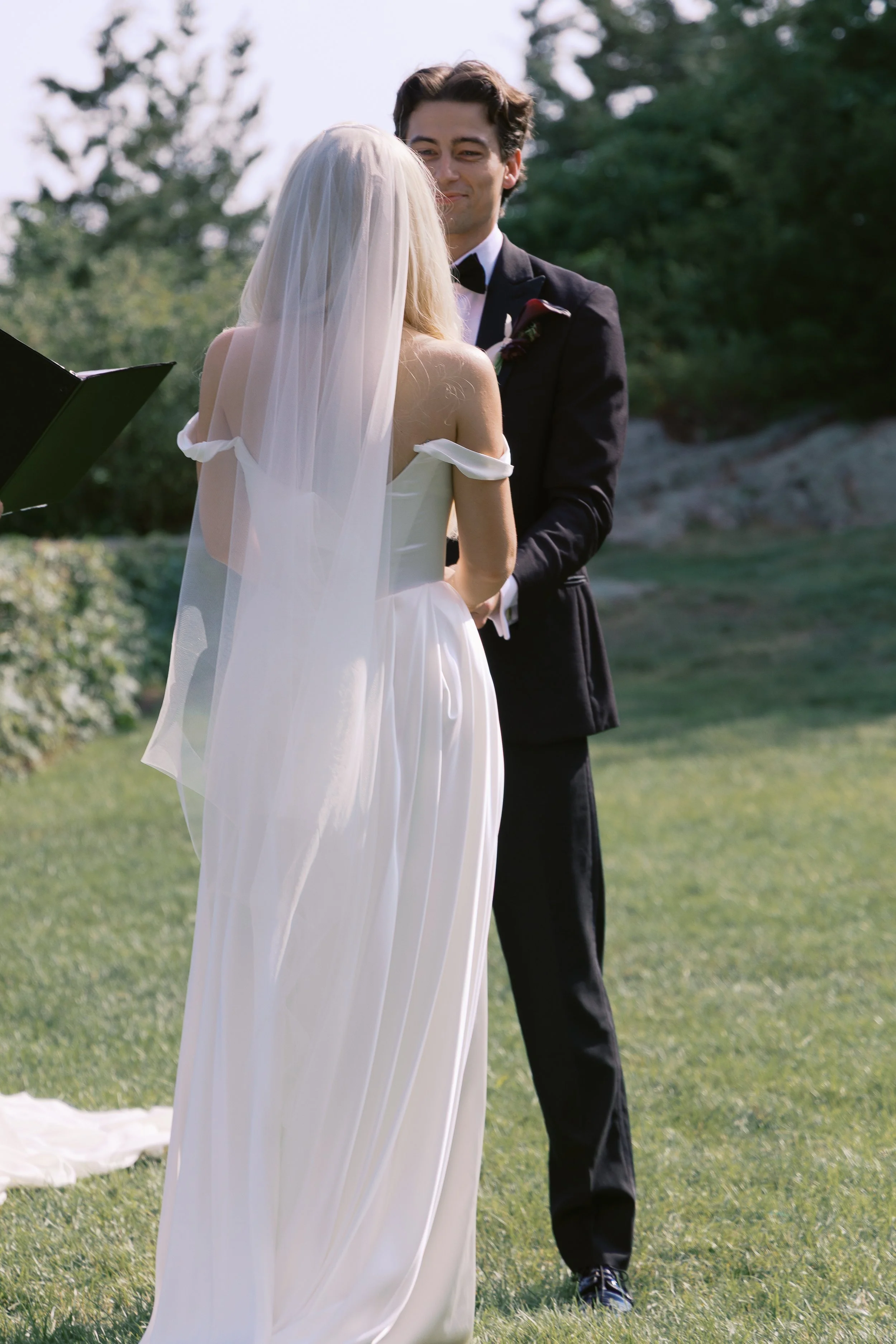 A couple getting married outdoors, with the bride in a white gown and veil and the groom in a black tuxedo, standing on grass in front of trees, during their wedding ceremony.