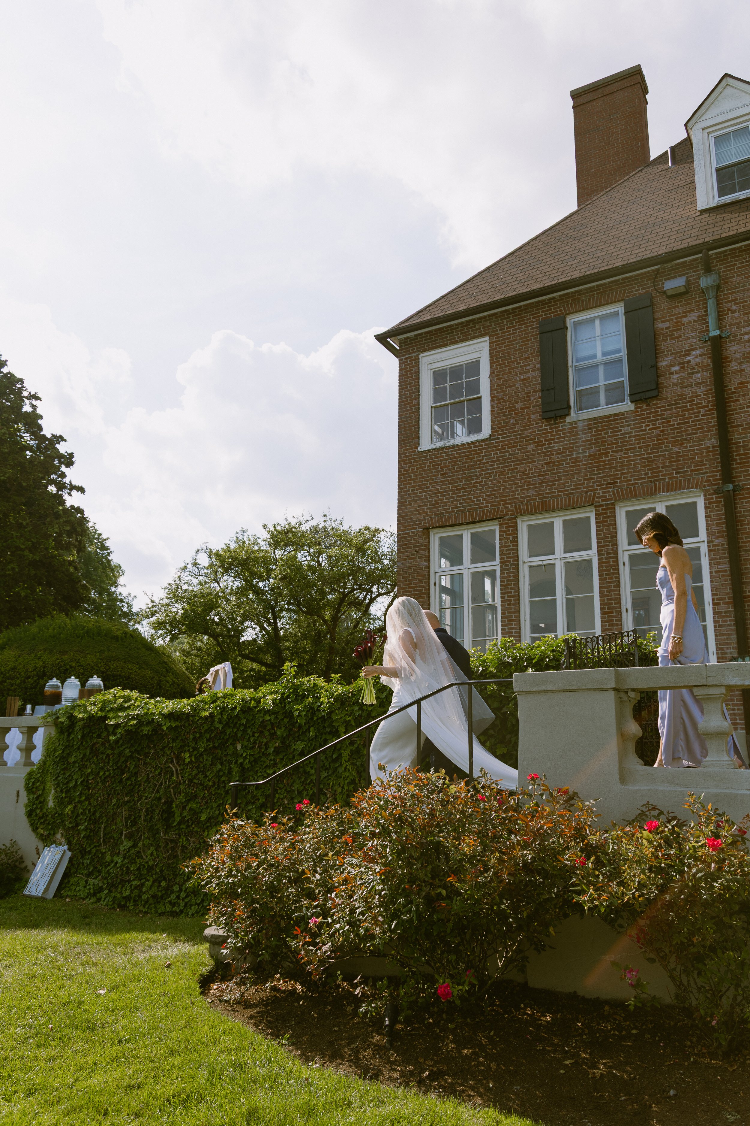 A bride in a white wedding dress and veil walking up the stairs of a large brick house, holding a bouquet of flowers. An officiant and a bridesmaid or guest are nearby on the stairs. The scene takes place outdoors on a sunny day with greenery and flo