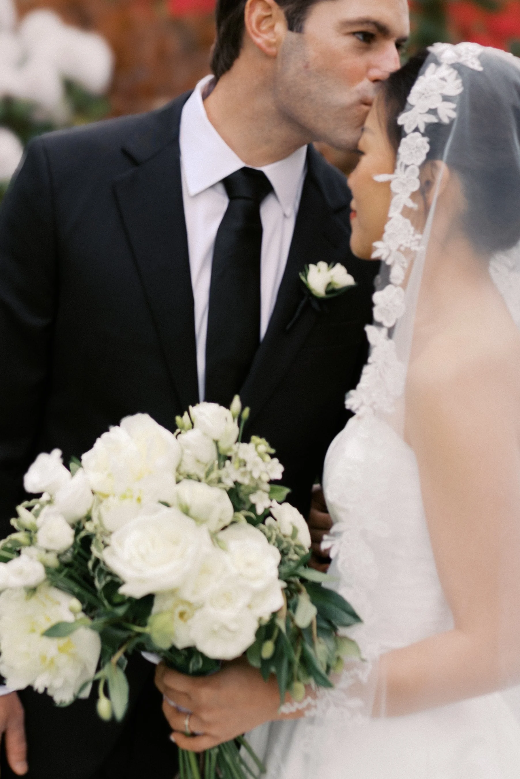A groom in a black suit and tie kisses a bride in a wedding dress and lace veil, holding a bouquet of white flowers.