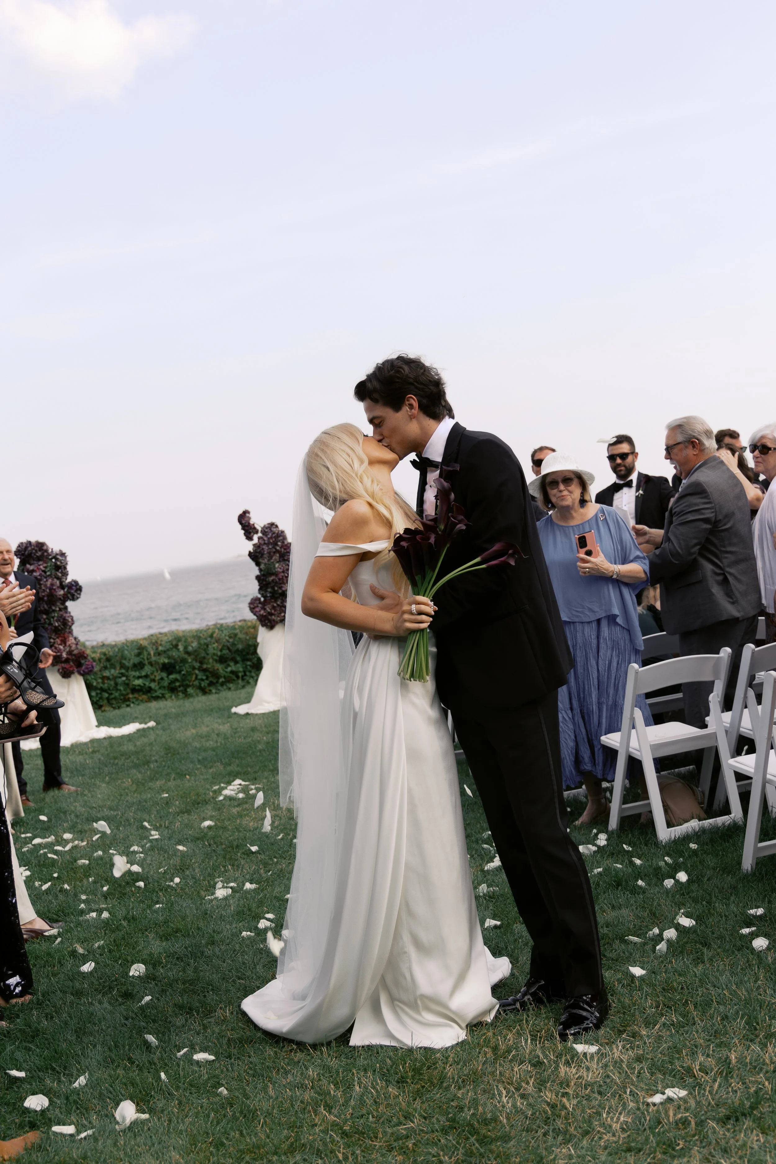 A newlywed couple sharing a kiss at an outdoor wedding ceremony by the ocean, with guests standing and sitting nearby.