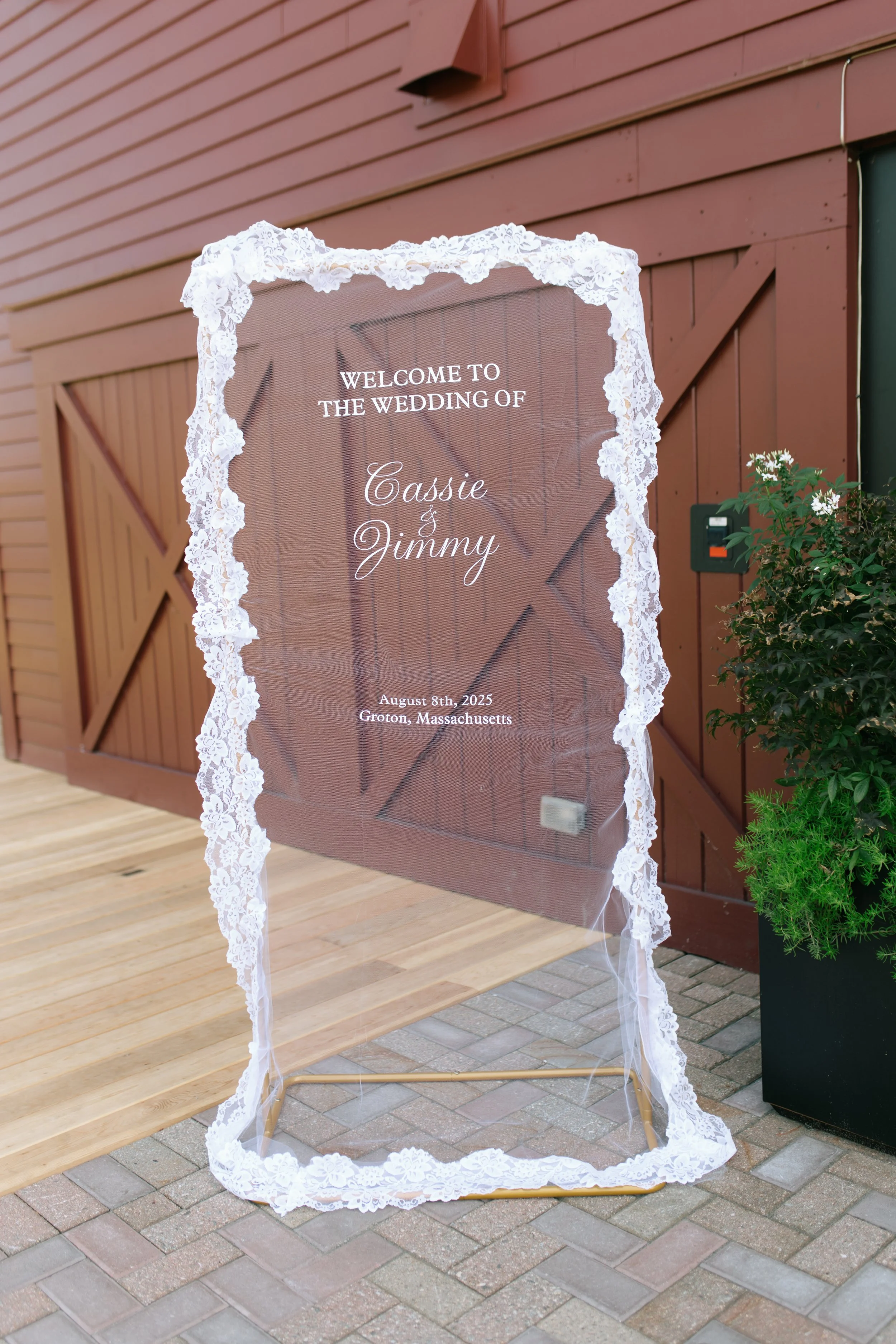 Wedding welcome sign in front of a barn with brown wooden siding, decorated with white lace and ribbon border, displaying the names Cassie and Jimmy, with the date August 8th, 2025, and location Groton, Massachusetts.