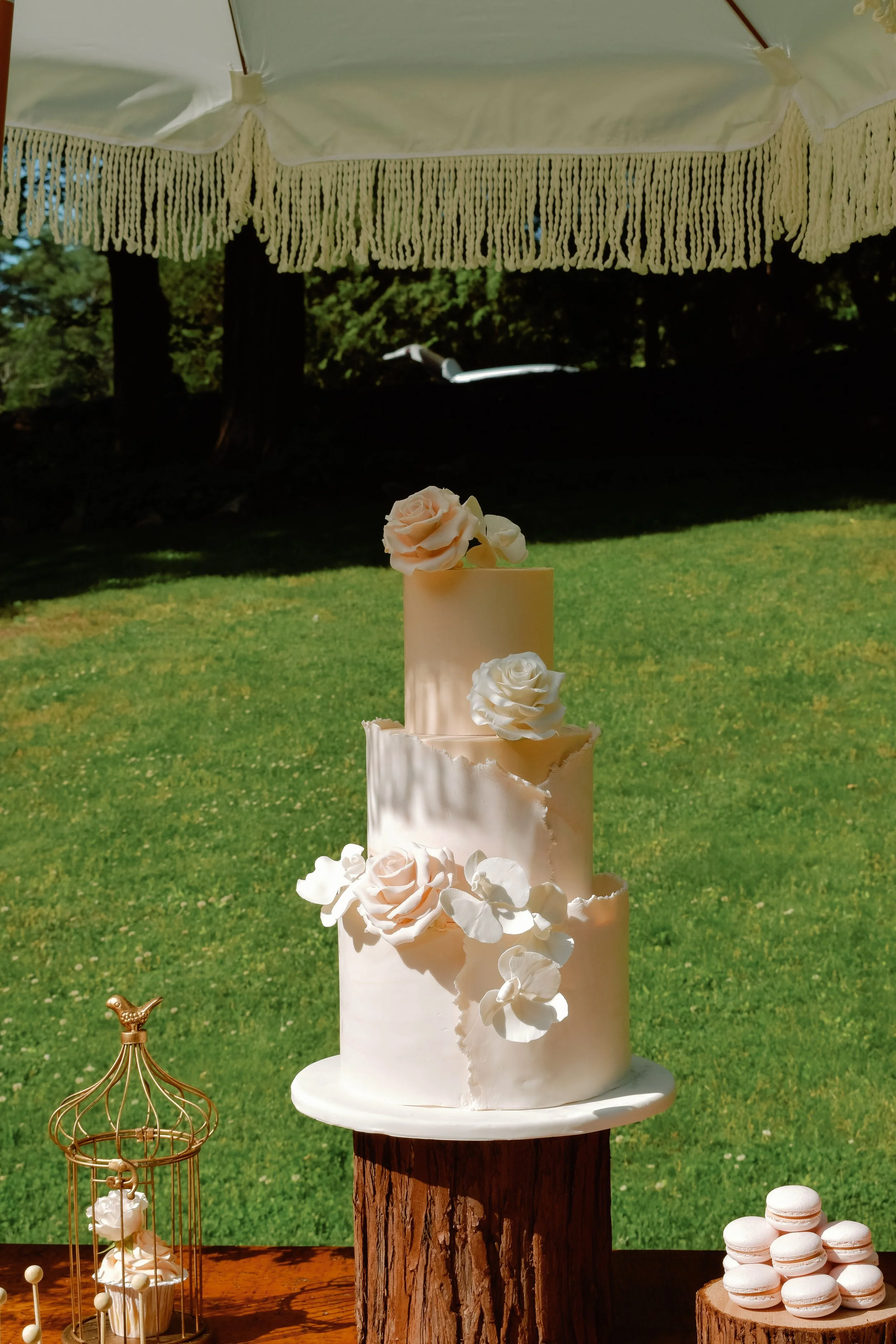Three-tiered white wedding cake decorated with white and blush roses and white orchids, set outdoors on a wooden table under a white fringed canopy, with macarons and a golden birdcage nearby.